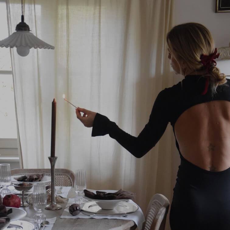 Žena v černých šatech s odhalenými zády zapaluje svíčku na elegantně prostřeném stole v jídelně
