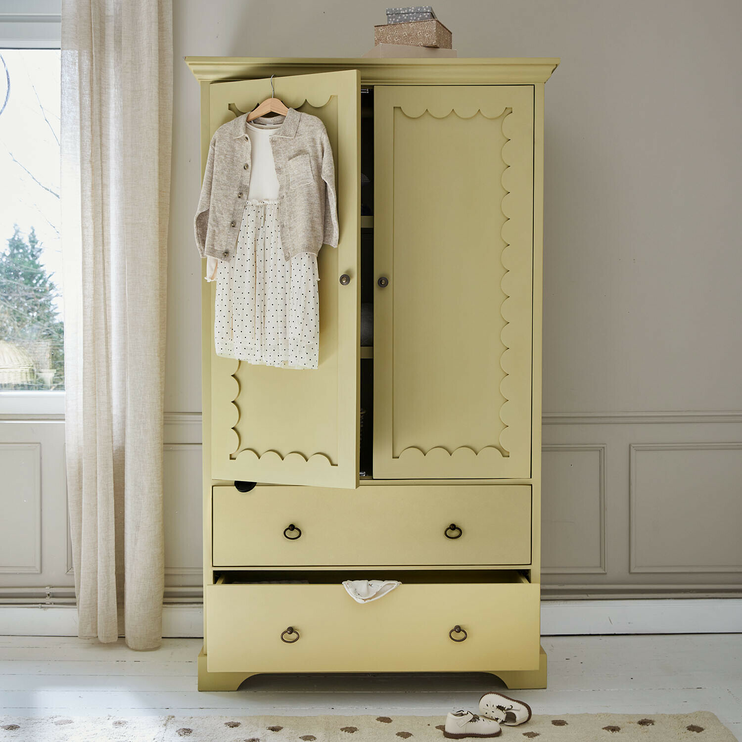 Dětská šatní skříň Frollin Žlutá dřevěná skříň s ozdobnými dveřmi a dětským oblečením zavěšeným na dveřích v světlé místnosti