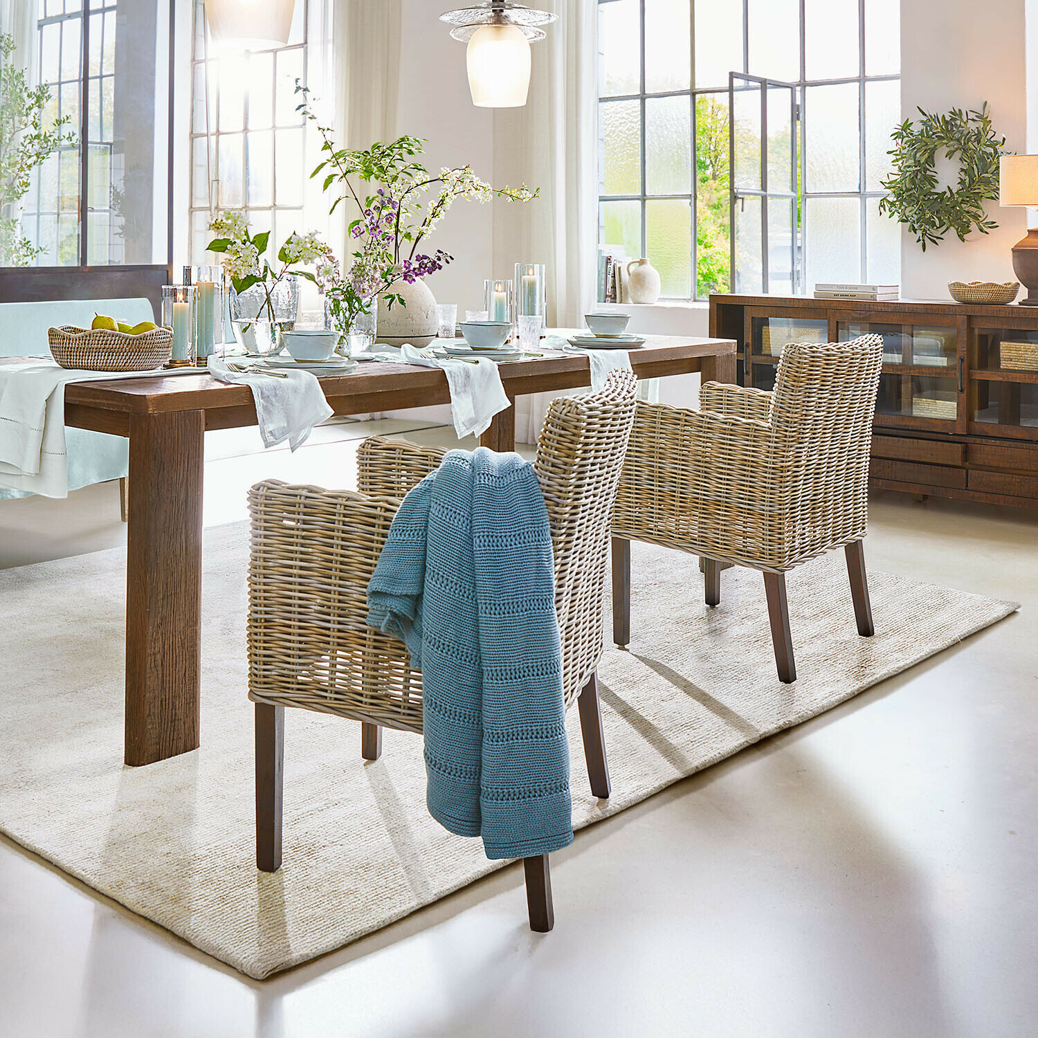 Křeslo Forêt Douce Jídelna s dřevěným stolem a přírodními ratanovými židlemi, květinová výzdoba a modrý pléd