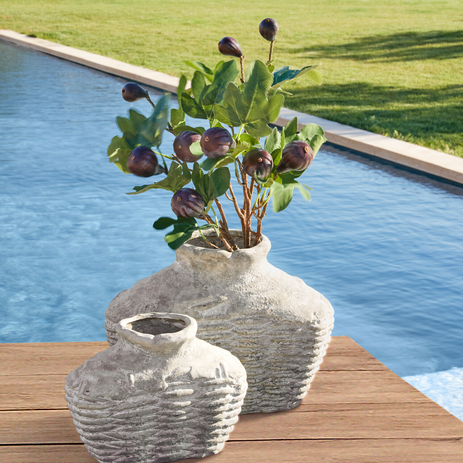 Dvě rustikální šedé keramické vázy na dřevěném stole u bazénu se zelenými listy a plody