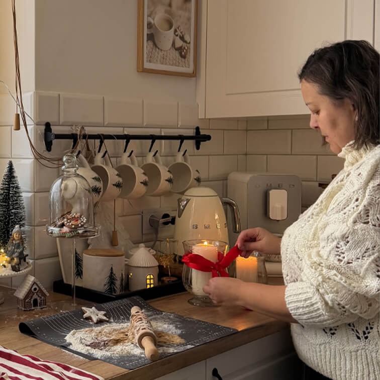 Žena v bílém svetru zdobí svíčku červenou stuhou v útulné kuchyni s pečícími pomůckami