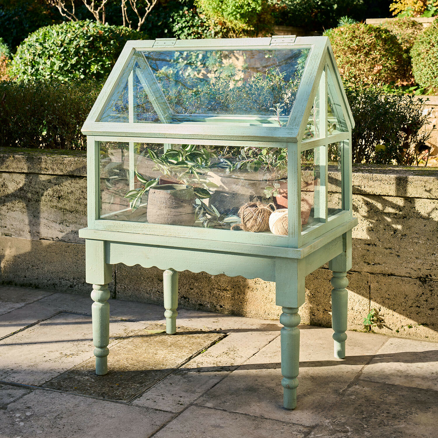 Malá zelená dřevěná skleník na nožkách s rostlinami a klubky provázku do zahrady