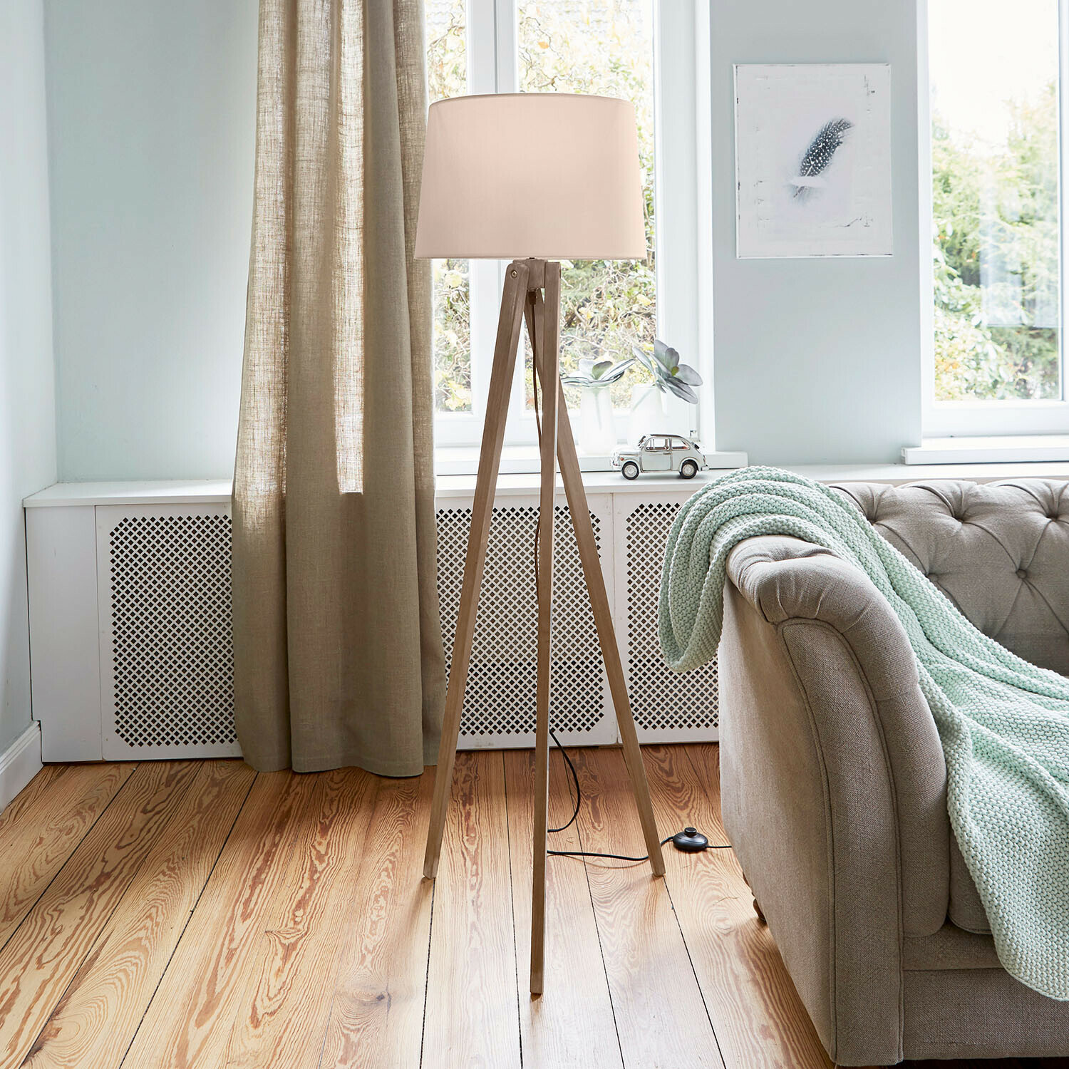 Stojací lampa s dřevěným stojanem a krémovým stínidlem v světlém obývacím pokoji s pohovkou a dekou