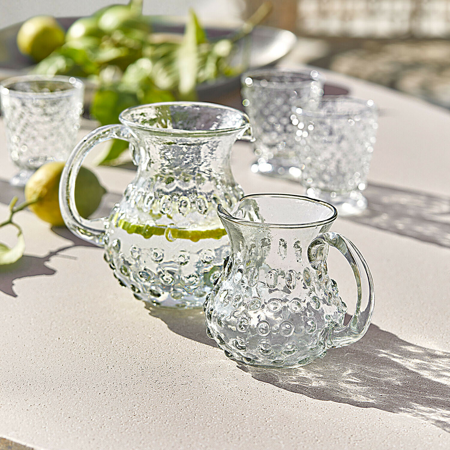 Skleněná karafa a džbánek s reliéfním vzorem na světlém stole, vhodné na letní nápoje