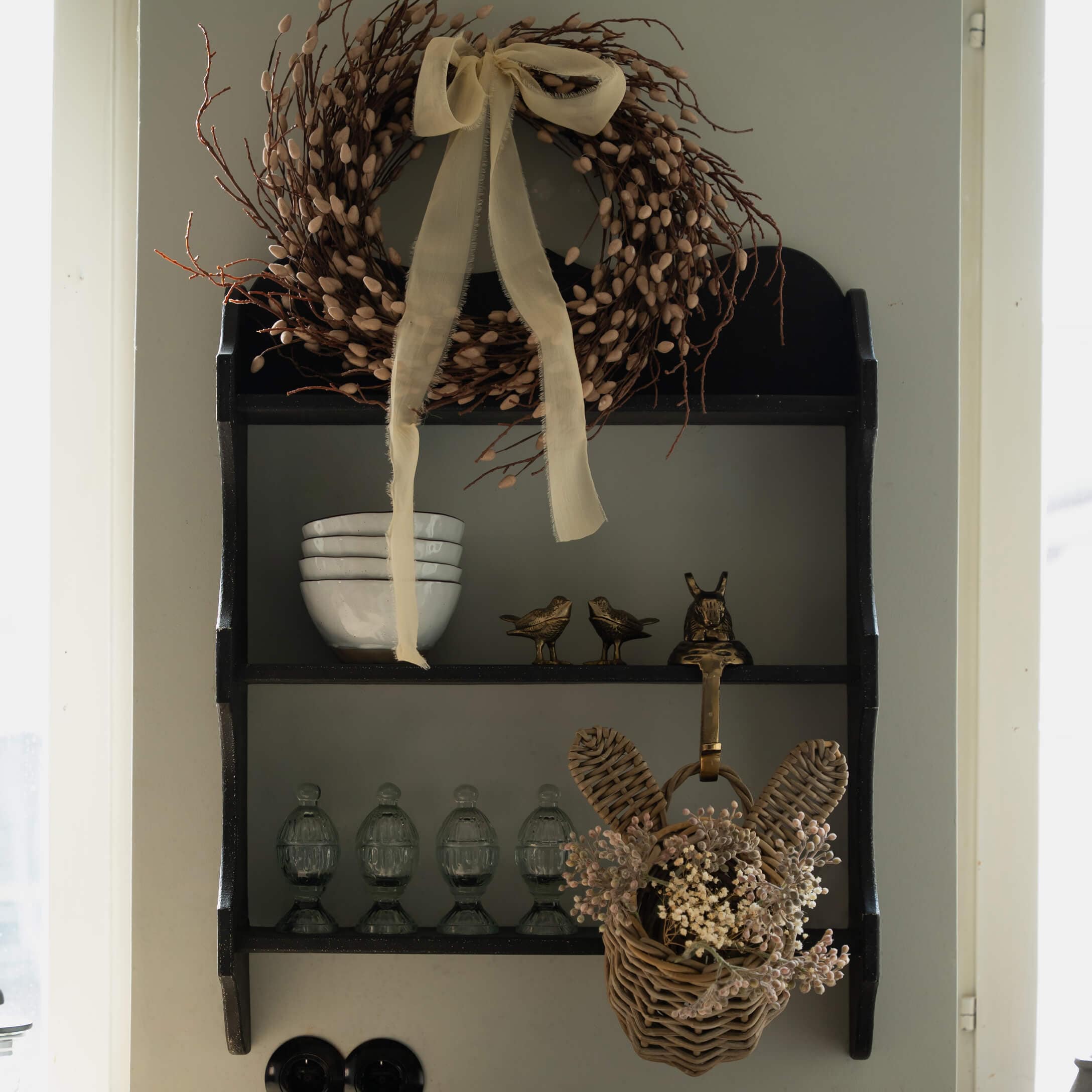 Černá dřevěná nástěnná police s dekorativním věncem a proutěným košíkem se sušenými květinami