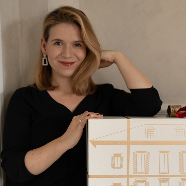 Žena v černém topu opírající se o bílou krabici s kresbou domu, interiér, dárek