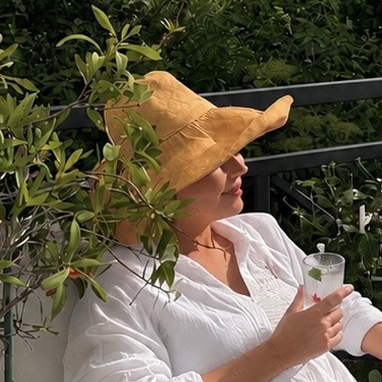 Žena v bílé košili a žlutém slaměném klobouku odpočívá na terase s nápojem v ruce