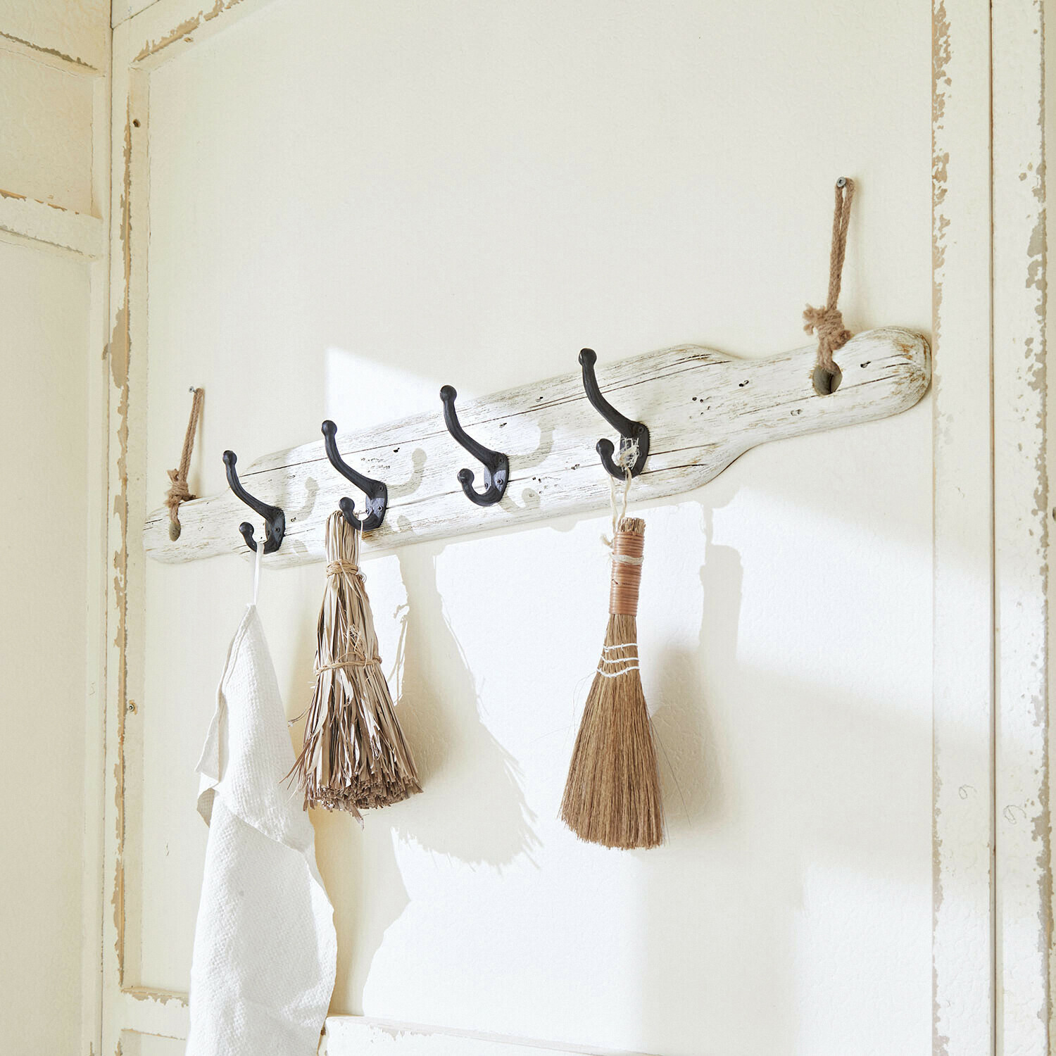 Rustikální dřevěný nástěnný věšák v bílé barvě s černými háčky a přírodními dekorativními košťaty