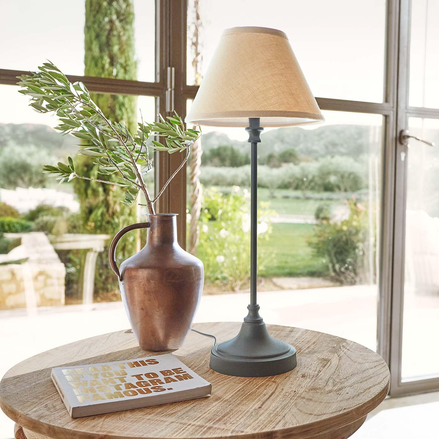 Měděný kovový džbán s větví a stolní lampa s béžovým stínidlem na dřevěném stole