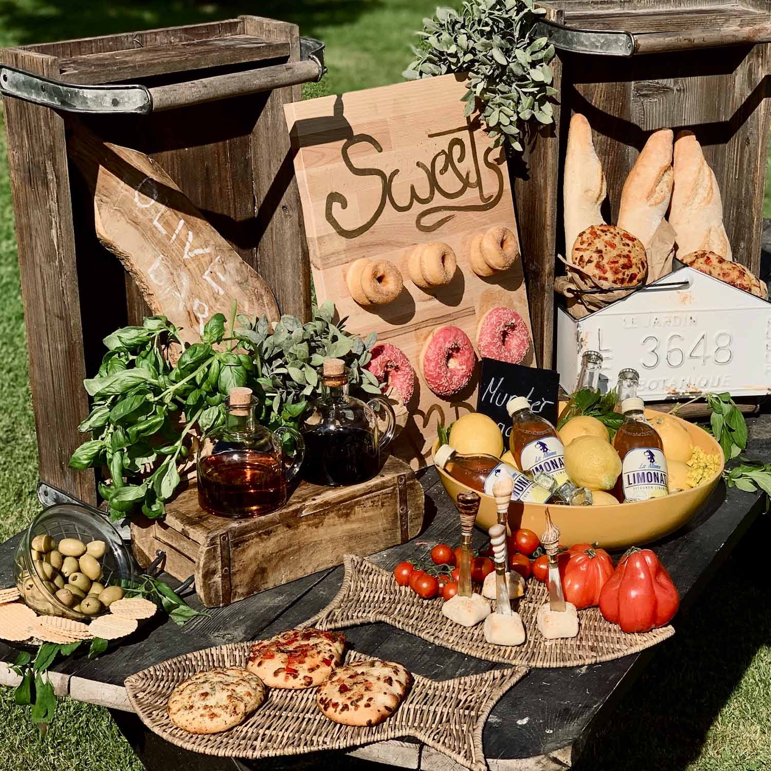 Rustikální stůl s dřevěnými bedýnkami, pečivem, olivami, bylinkami a limonádou venku na slunci