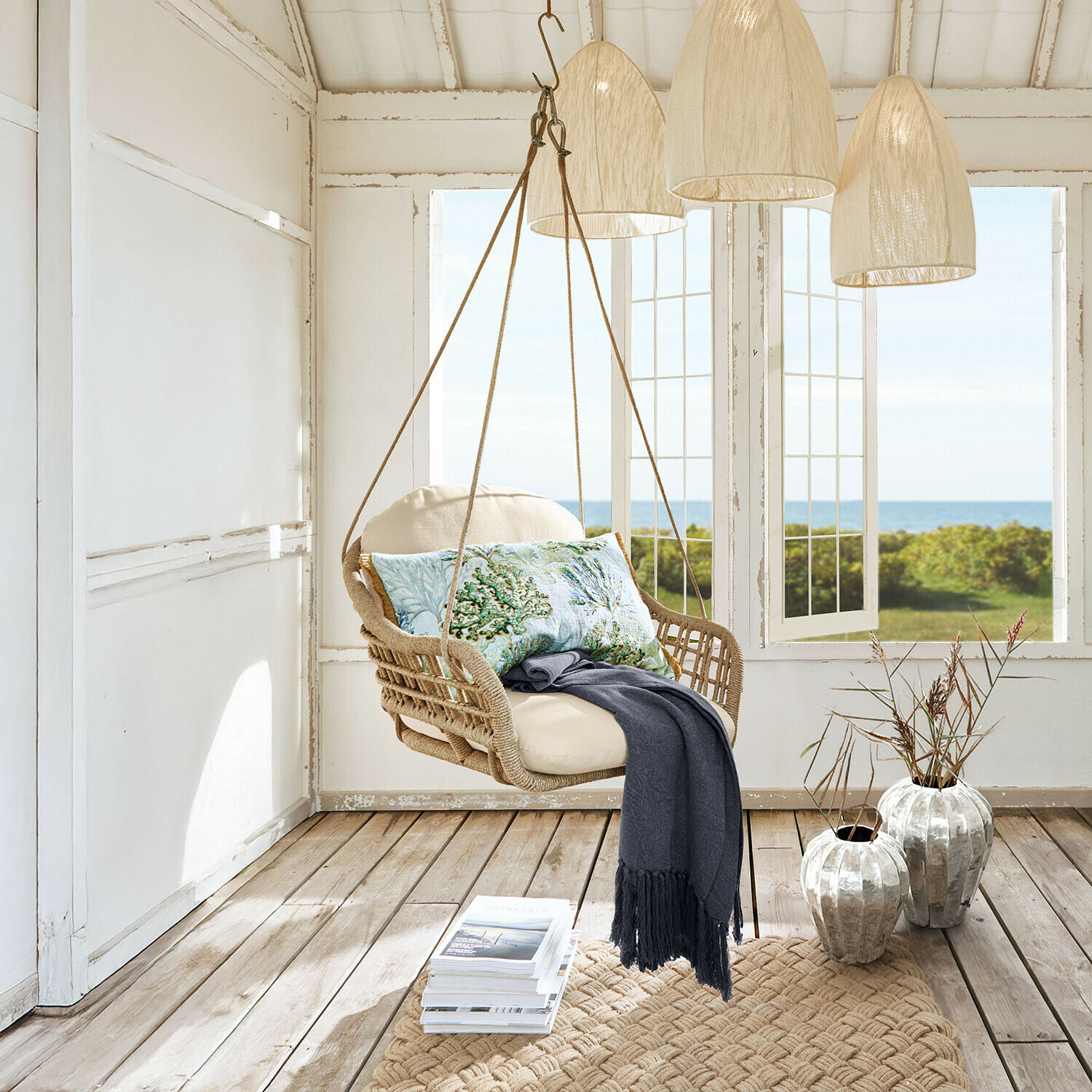Závěsné křeslo z pleteného lana s krémovým polstrováním a dekorativním polštářem s mořským motivem v světlé místnosti