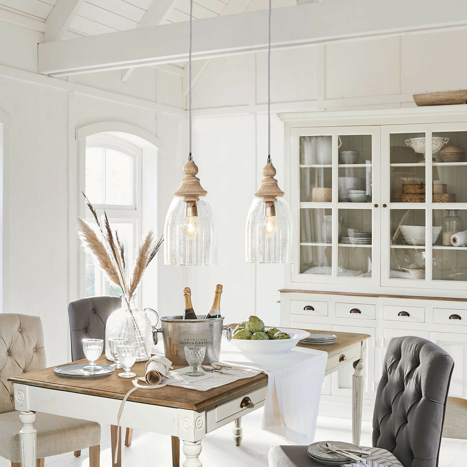 Rustikální jídelna s bílým dřevěným stolem, šedými čalouněnými židlemi a vitrínou s dekorem z proutí