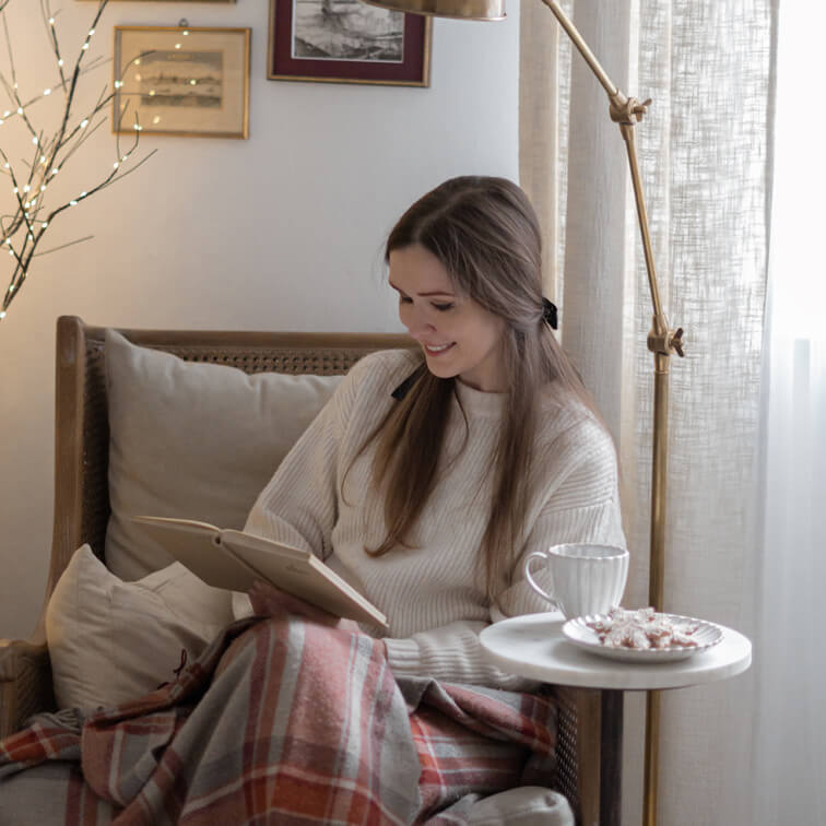 Žena čtoucí knihu v proutěném křesle s krémovým svetrem a teplou kostkovanou dekou