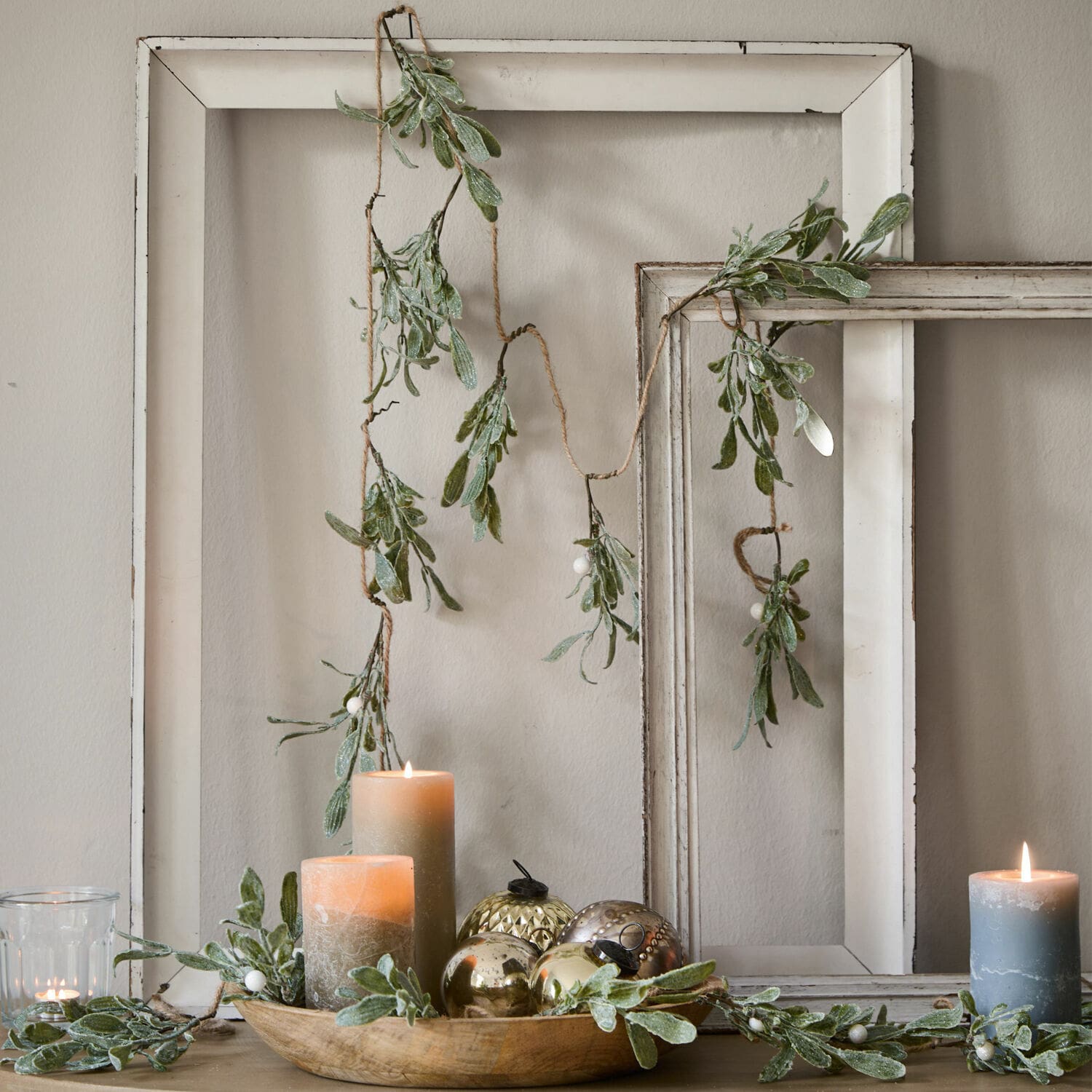 Girlanda Mistletoe Dekorativní girlanda se zelenými listy a bílými bobulemi na pozadí bílých dřevěných rámů, svíčky a ozdoby