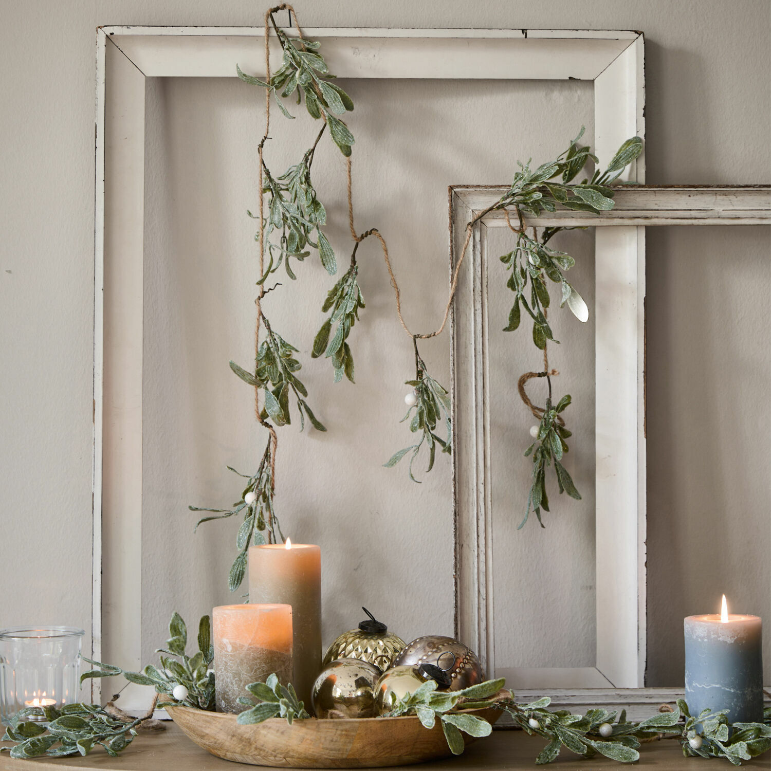 Girlanda Mistletoe Dekorativní girlanda se zelenými listy a bílými bobulemi na pozadí bílých dřevěných rámů, svíčky a ozdoby