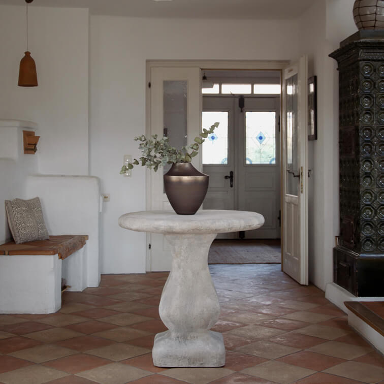 Kulatý světle šedý betonový stůl s tmavou keramickou vázi a zelenými listy v světlém interiéru