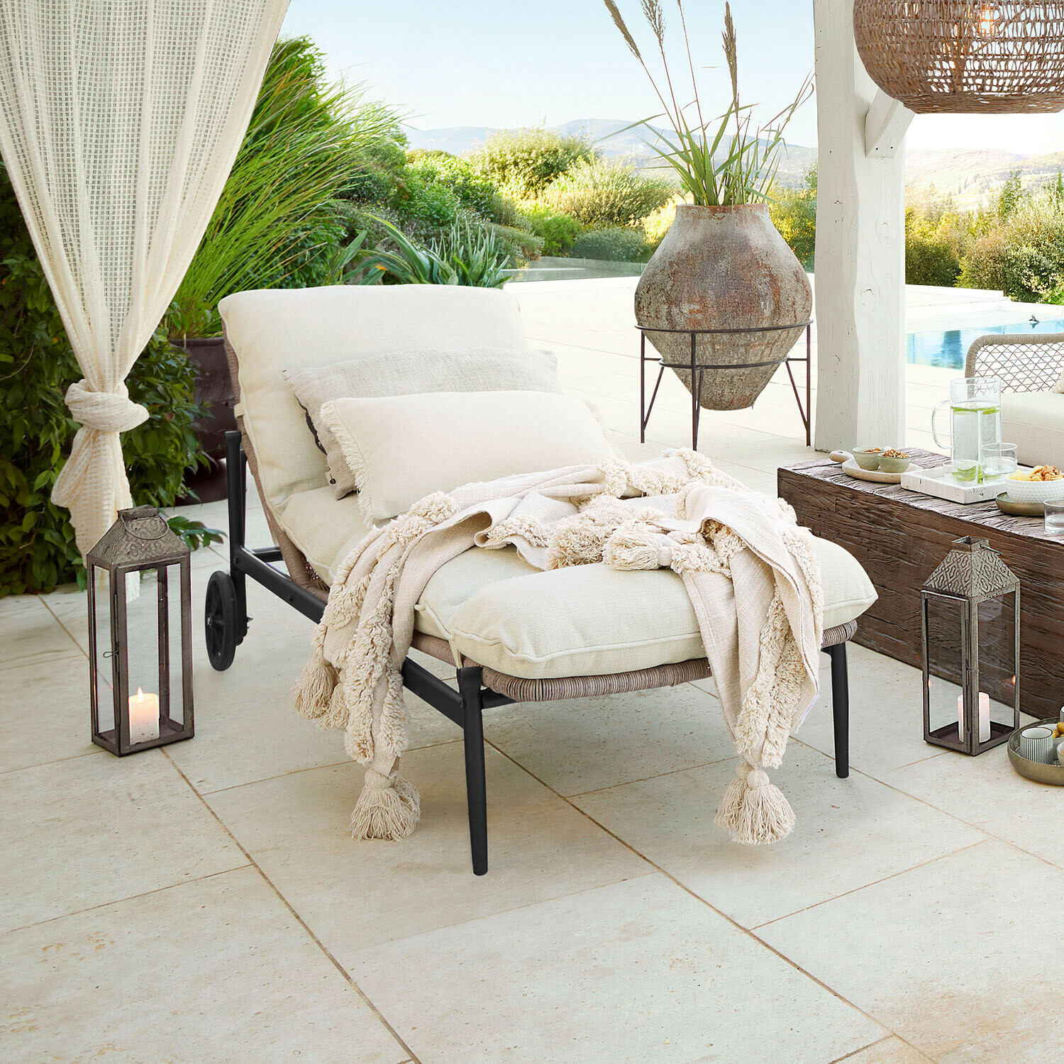 Zahradní lehátko s krémovými polštáři a měkkou dekou na terase s výhledem na bazén a zeleň