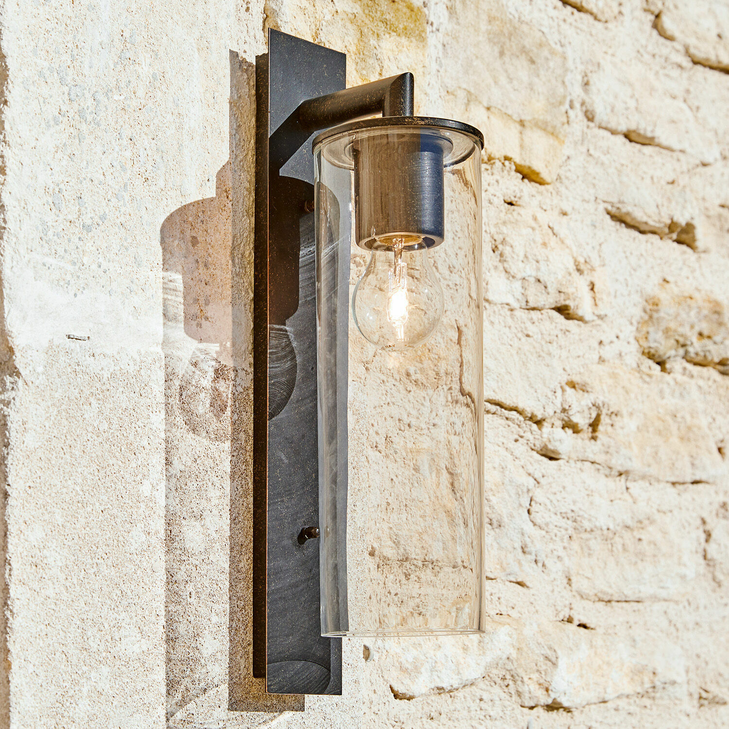 Venkovní nástěnná lampa Clisson Černé kovové nástěnné svítidlo s průhledným skleněným krytem na světlé kamenné zdi