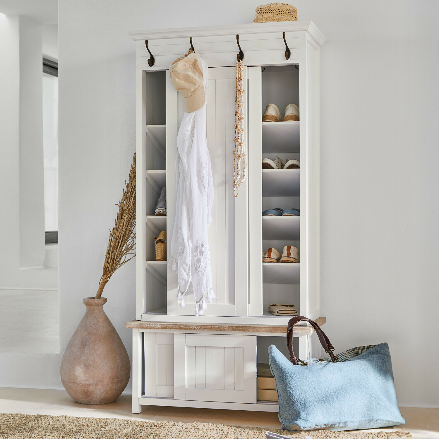 Bílá dřevěná botník s háčky a policemi, modrá taška a slaměný klobouk