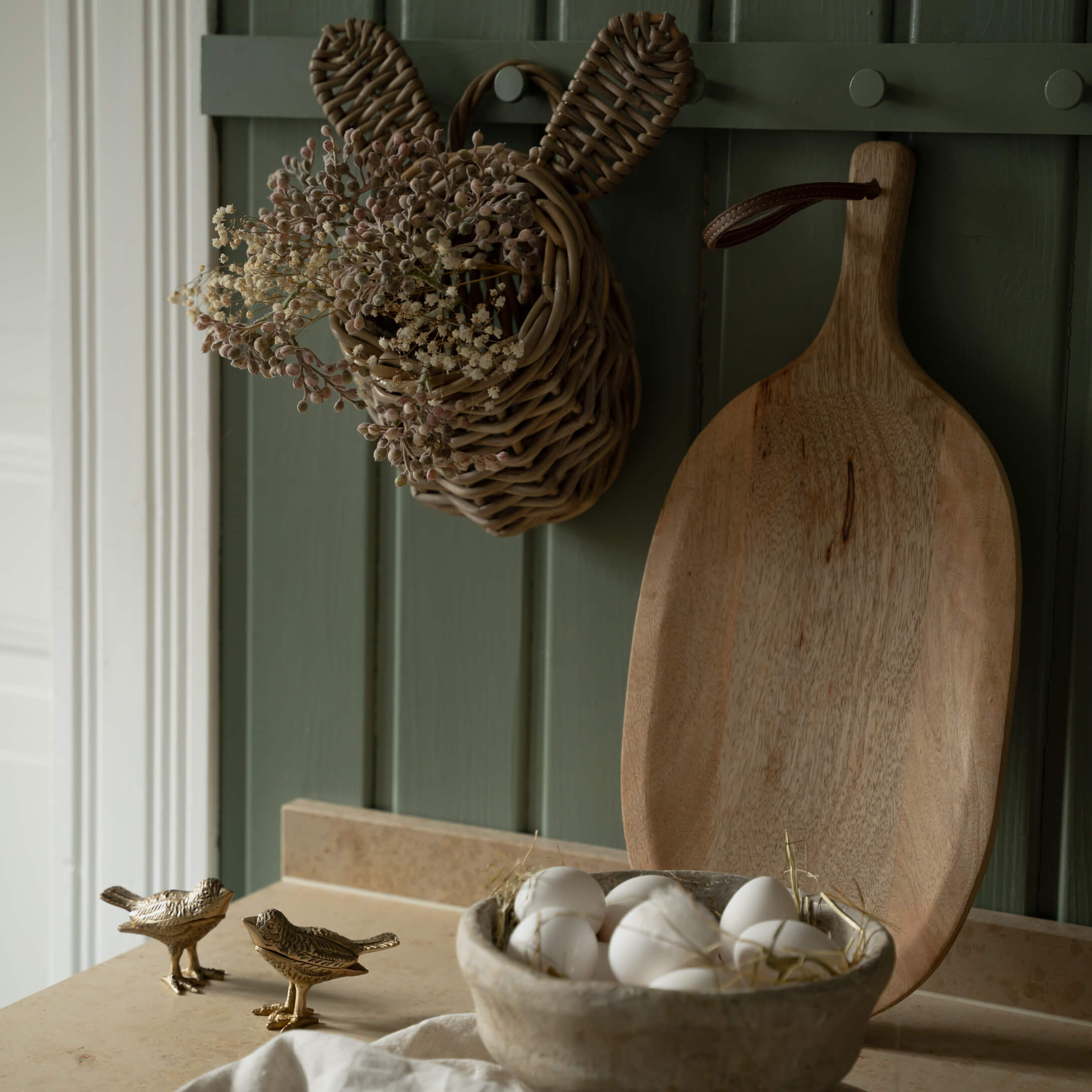 Proutěný košík ve tvaru králíka se sušenými květinami a dřevěná krájecí deska na zeleném pozadí