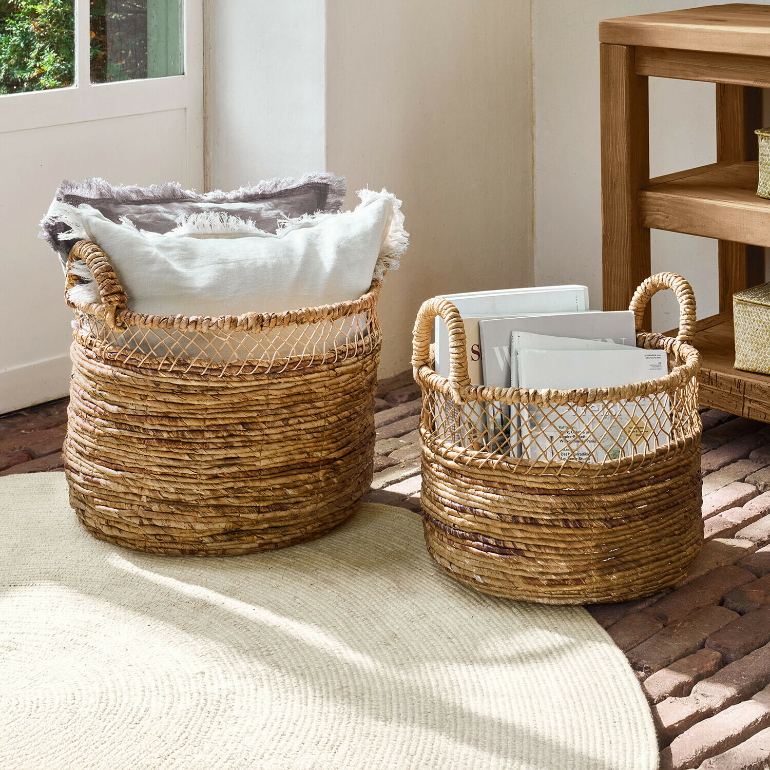 Dva přírodní proutěné koše s uchy, naplněné polštáři a knihami pro dekorativní úložný prostor
