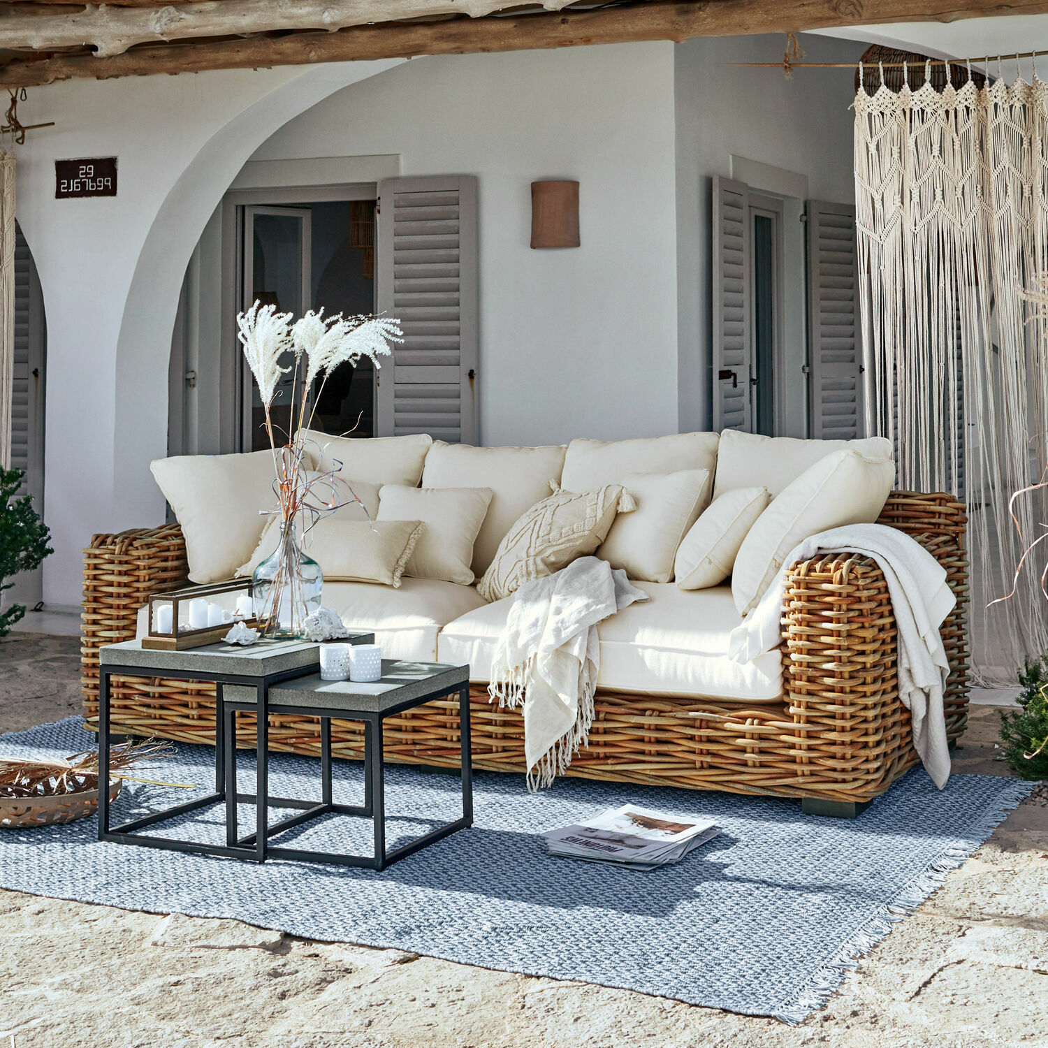 Pohovka Rimini Ratanová pohovka s krémovými polštáři a dekou na modrém koberci na terase