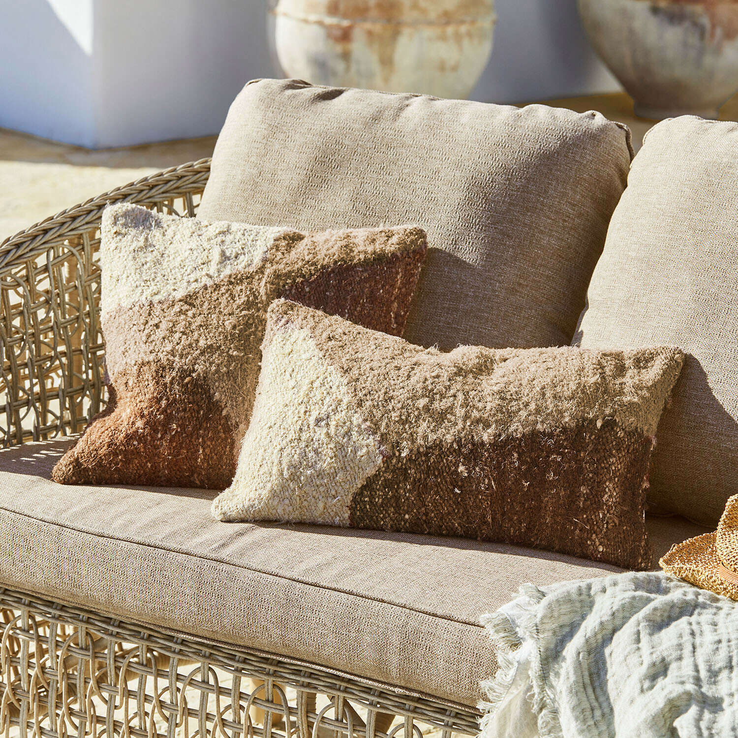 Béžová ratanová pohovka s měkkými polštáři v hnědých a krémových odstínech, dekorativní textilní deka