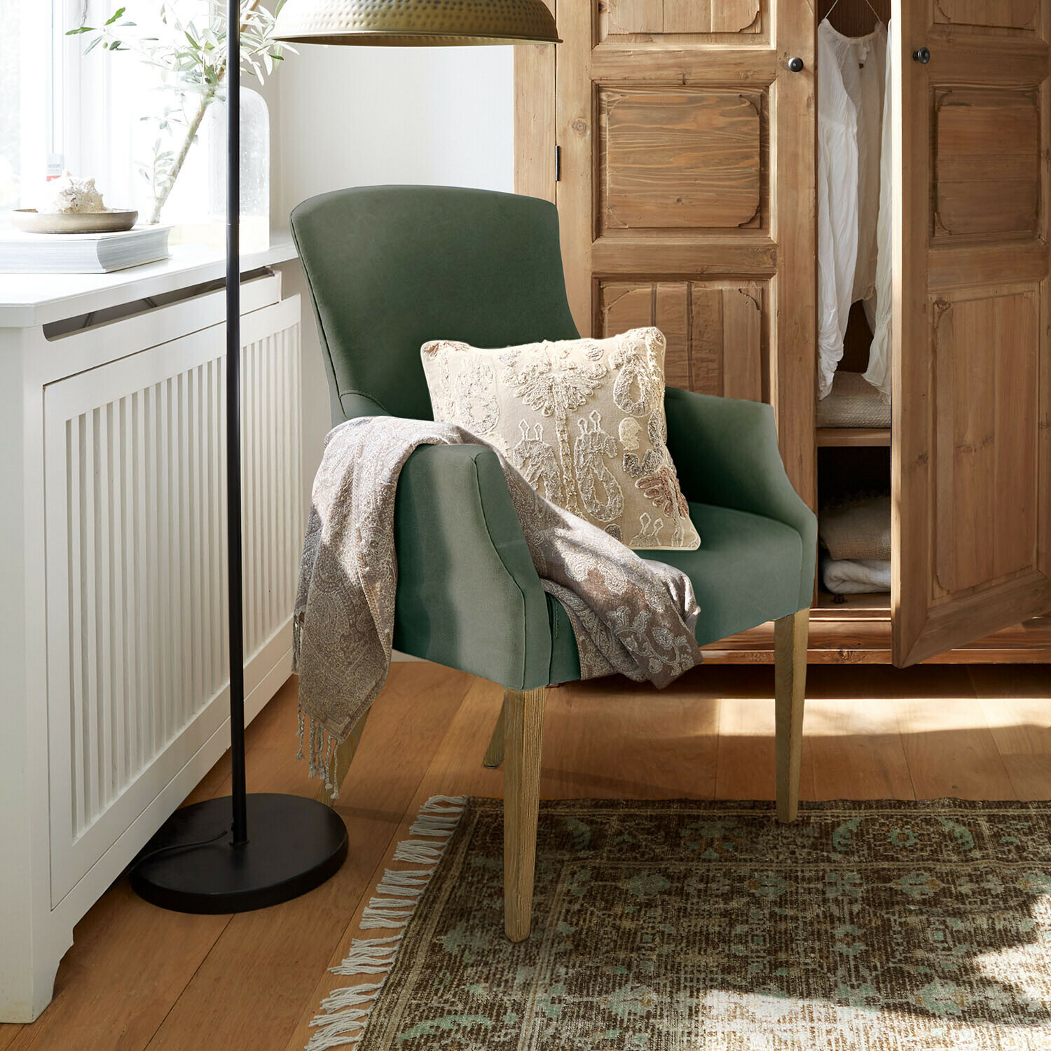 Zelené křeslo s dřevěnými nohami, krémový polštář a šedý pléd v útulném interiéru s dřevěnou skříní