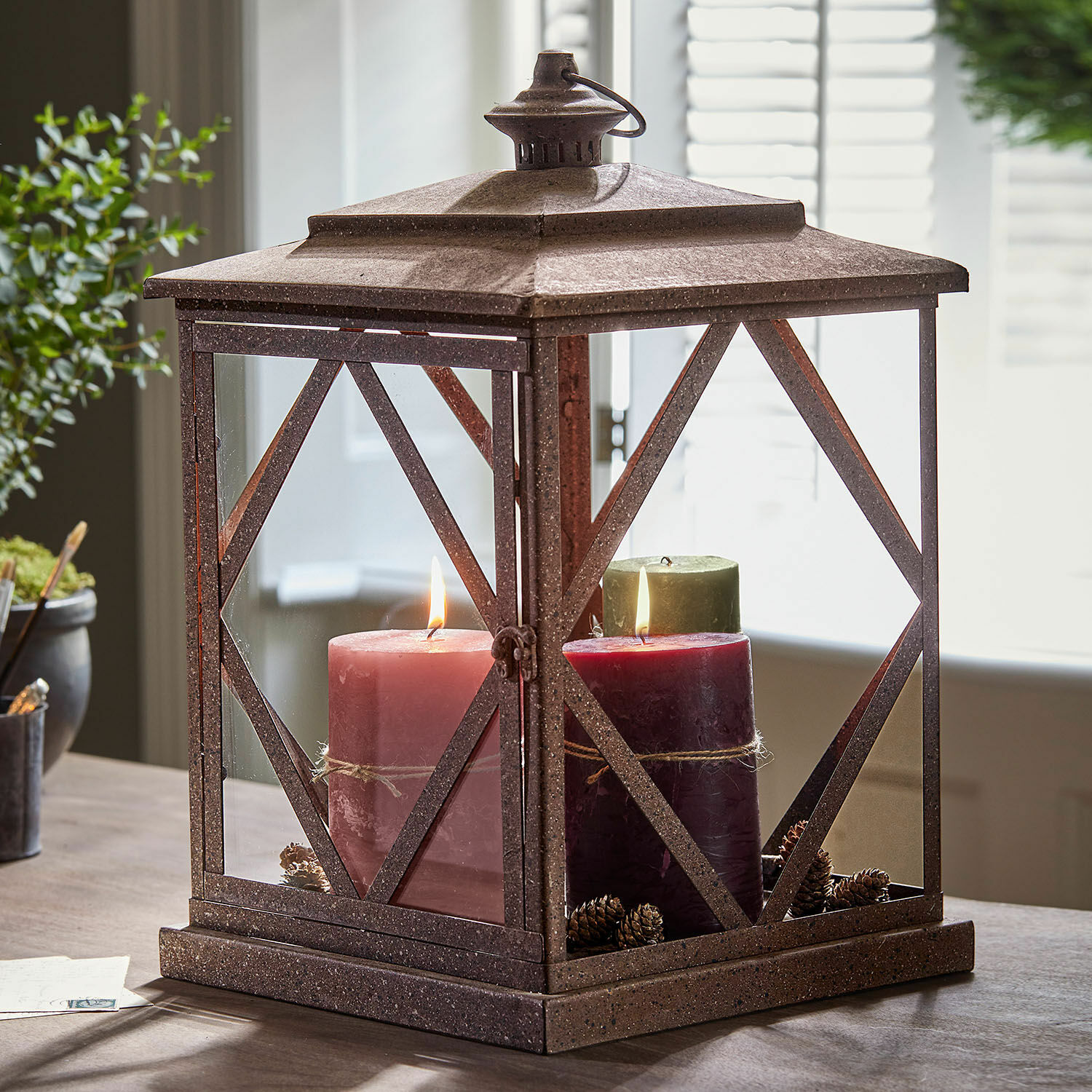 Dekorativní kovová lucerna rezavé barvy se třemi svíčkami a šiškami na stole