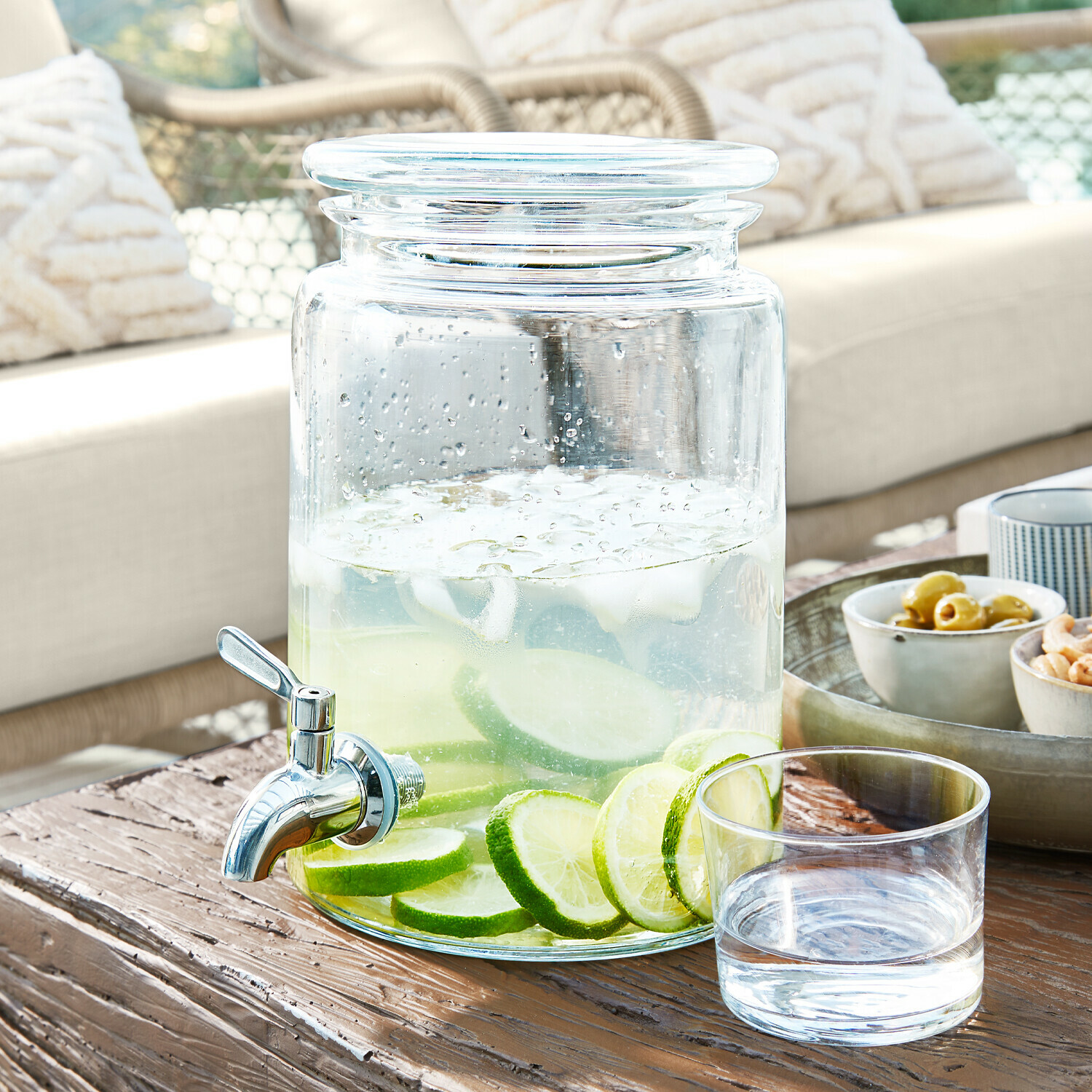 Skleněný nápojový dávkovač s kohoutkem, naplněný vodou s plátky limetky na dřevěném stole