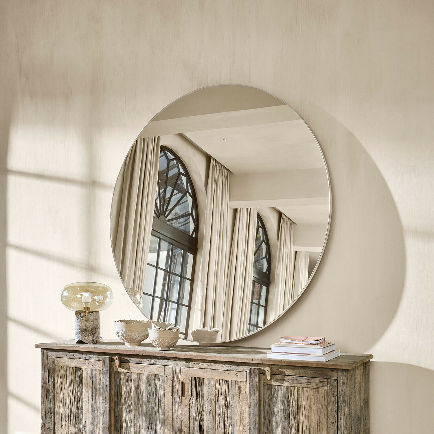 Velké kulaté zrcadlo na stěně nad dřevěnou komodou v světlém přírodním interiéru