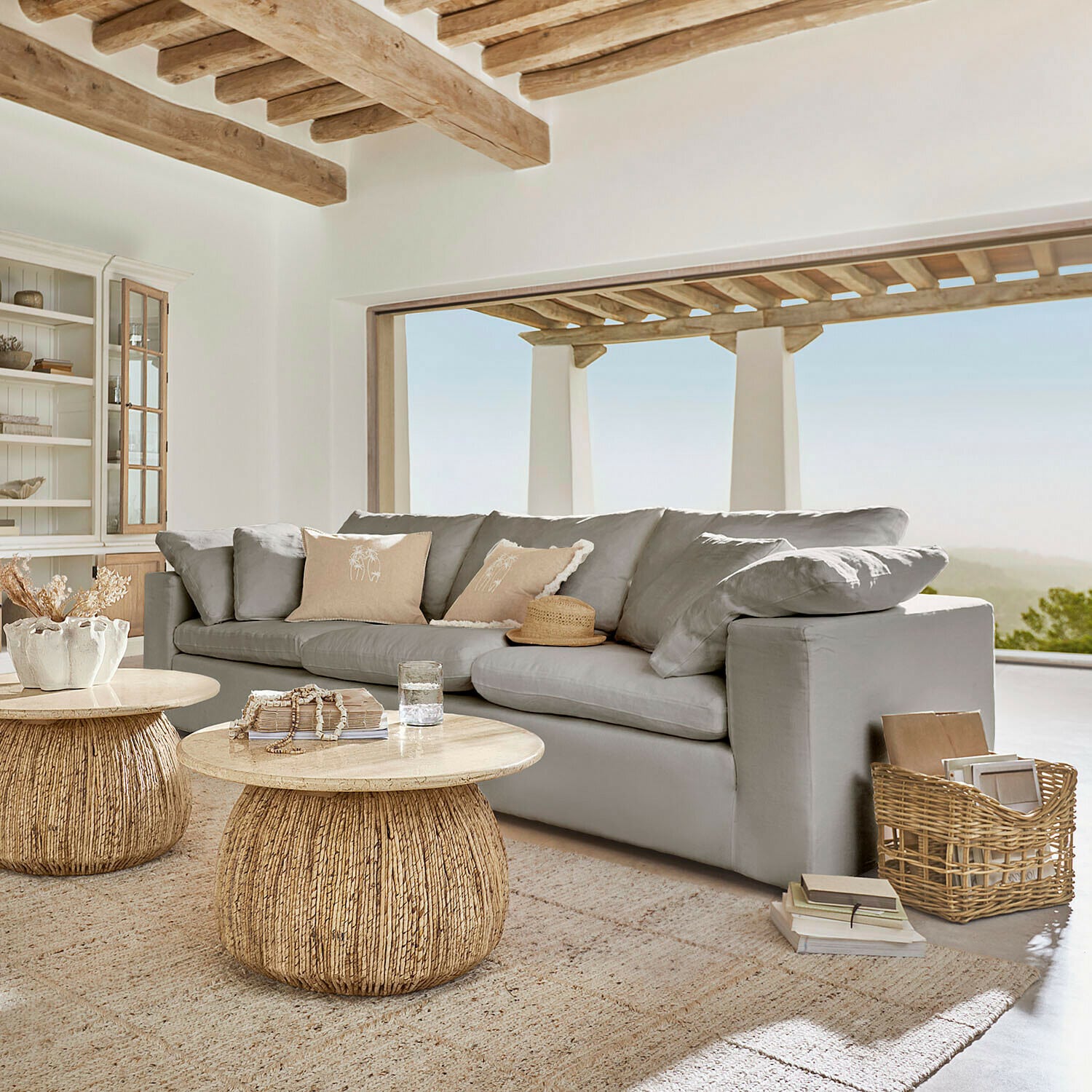 Šedá pohovka s měkkými polštáři a přírodními dřevěnými konferenčními stolky v světlém obývacím pokoji s výhledem