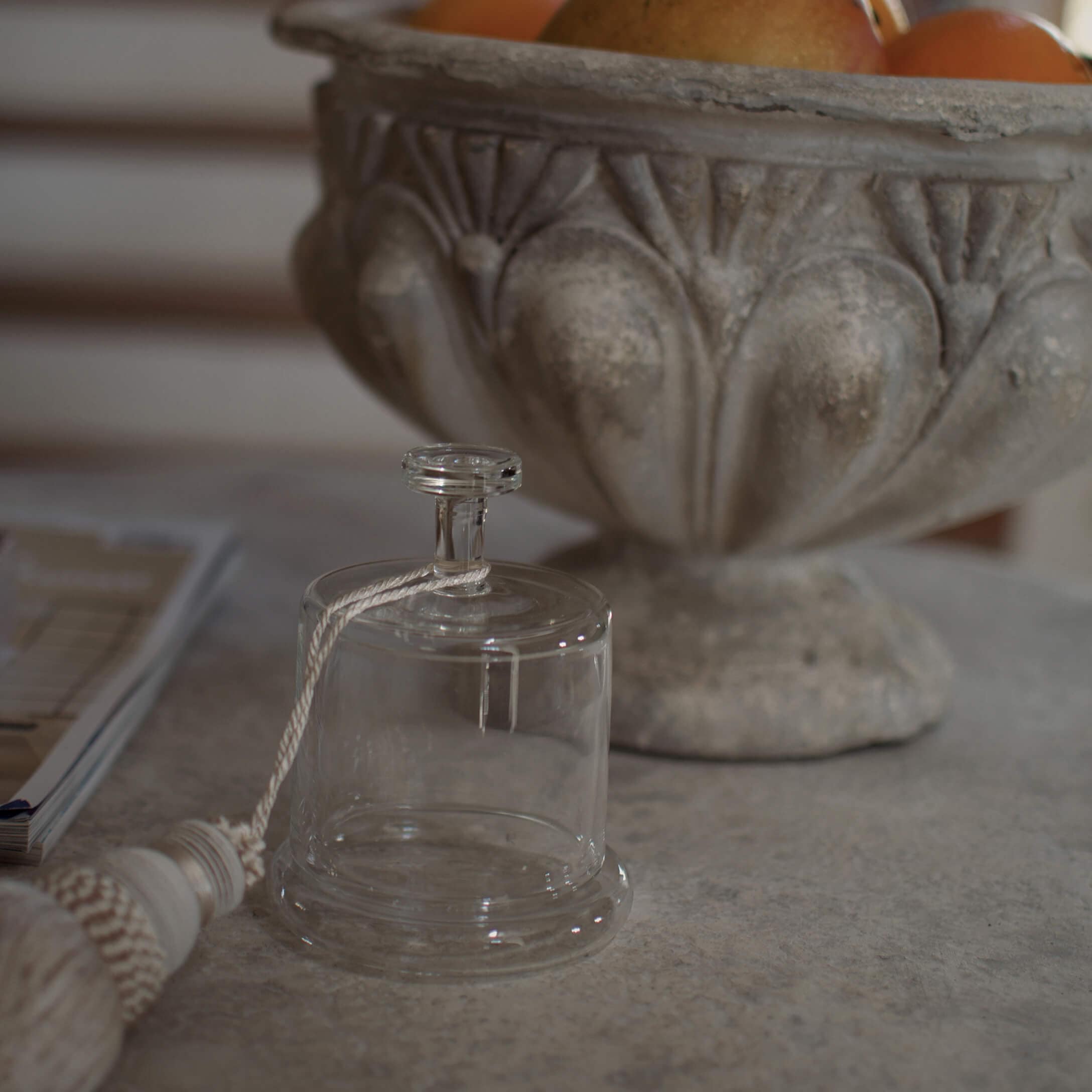 Průhledný skleněný zvonek s bílým provázkem na kamenném stole vedle zdobené mísy