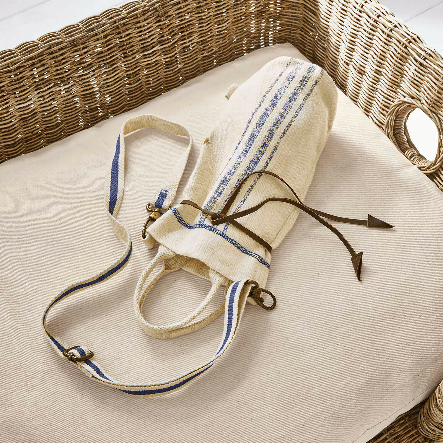 Béžová bavlněná taška s modrými pruhy a koženým šňůrkem na světlém polštáři s proutěným opěradlem