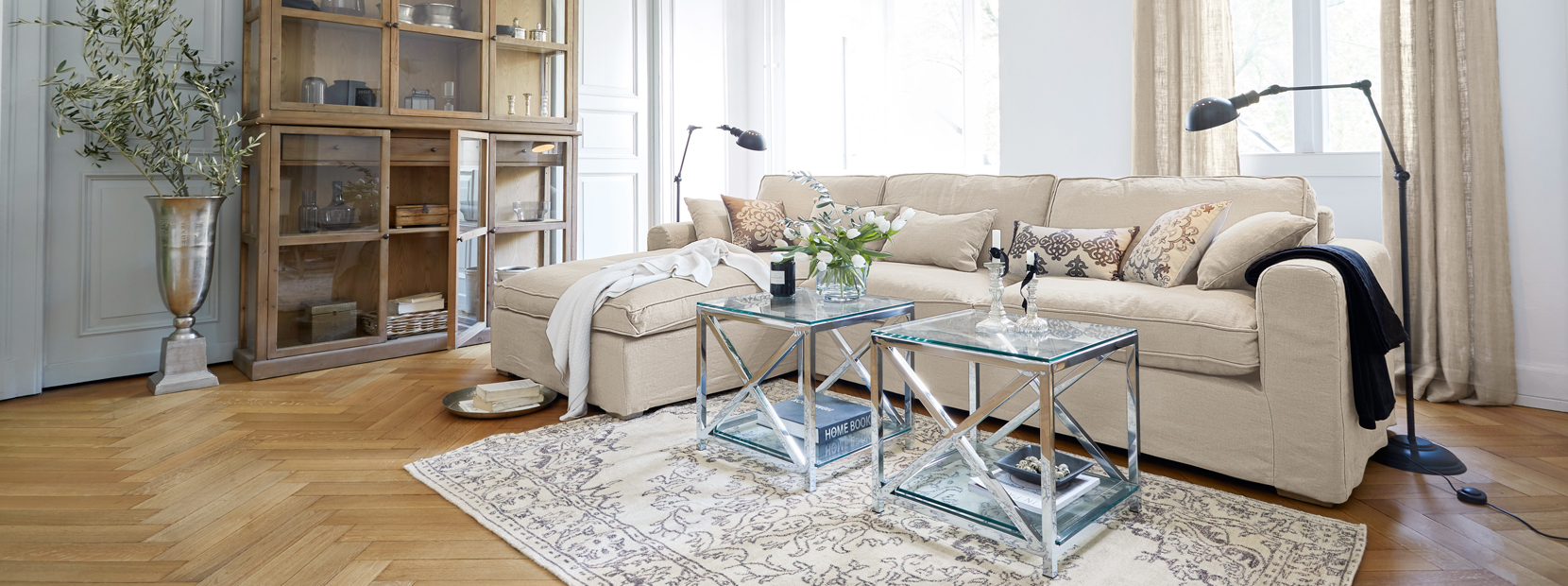 Klasický elegantní obývací pokoj s béžovou pohovkou, skleněnými stolky a dřevěnou vitrínou na světlé podlaze