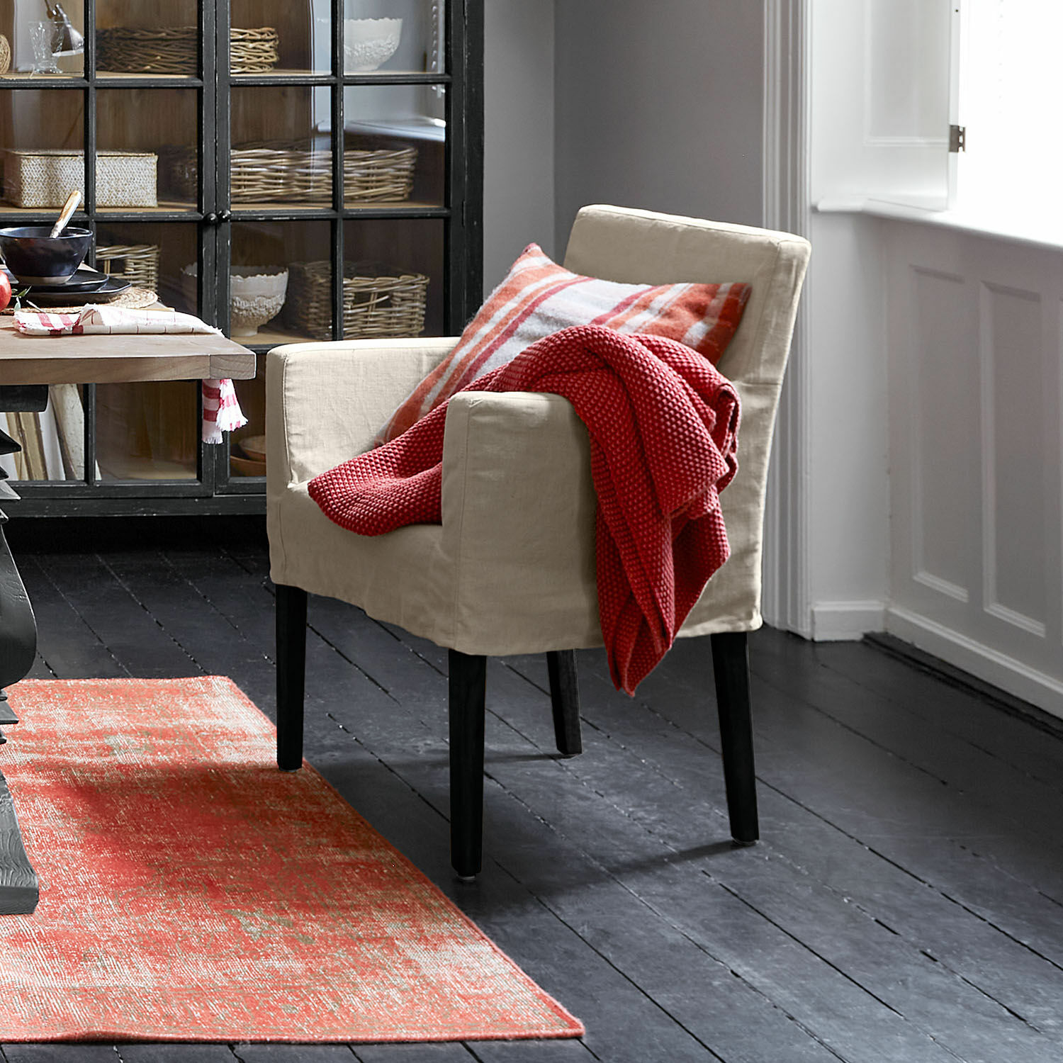 Béžové látkové křeslo s černými nohami, červená deka a kostkovaný polštář, červený koberec