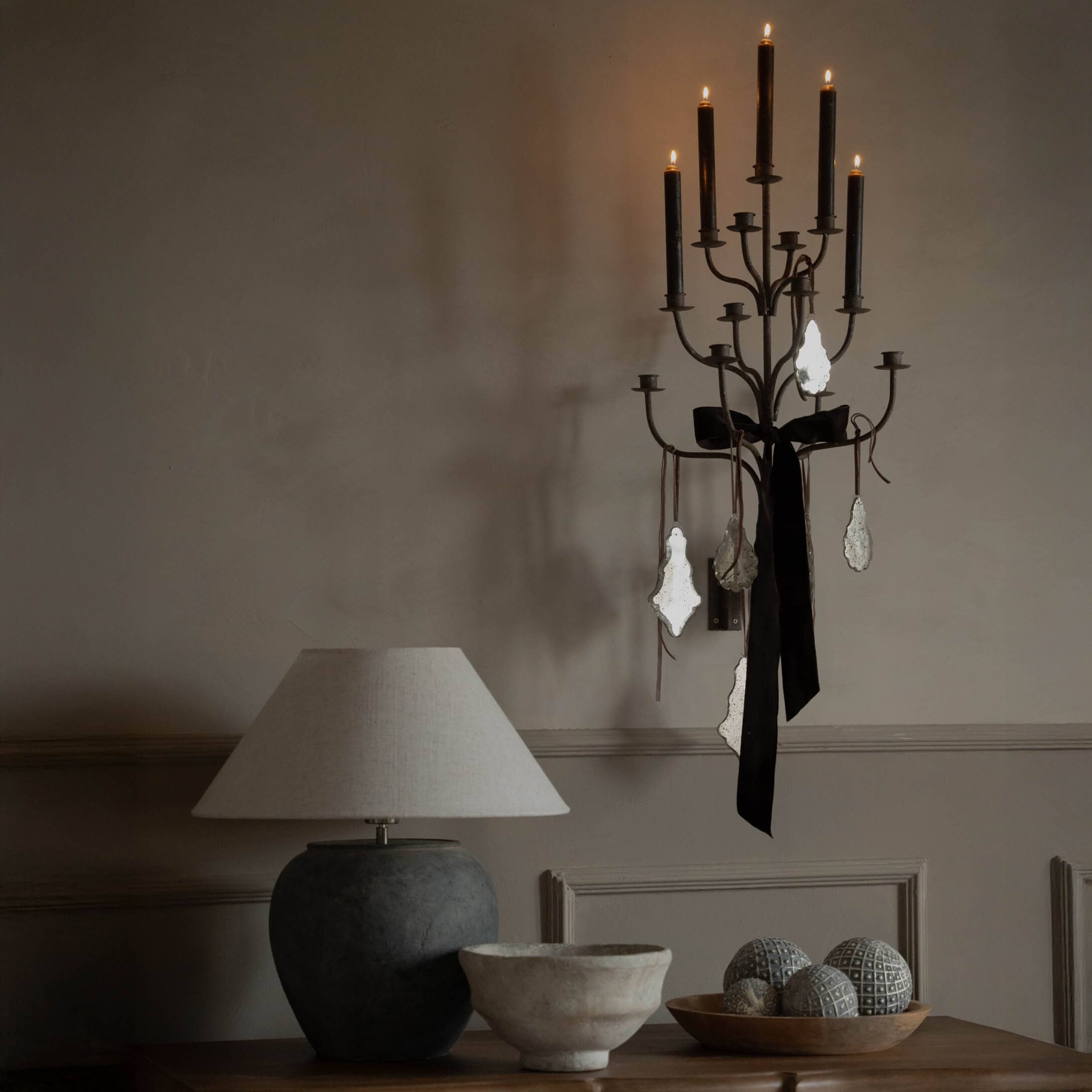 Černý kovový nástěnný svícen s černou mašlí a ozdobami, béžová stolní lampa na dřevěném stole