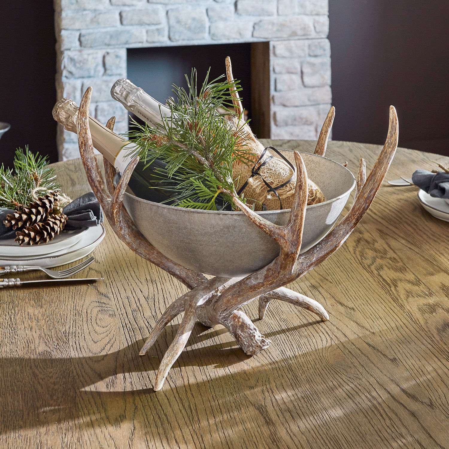 Dekorativní šedá keramická mísa na stojanu z paroží s lahvemi a zelenými větvičkami na dřevěném stole