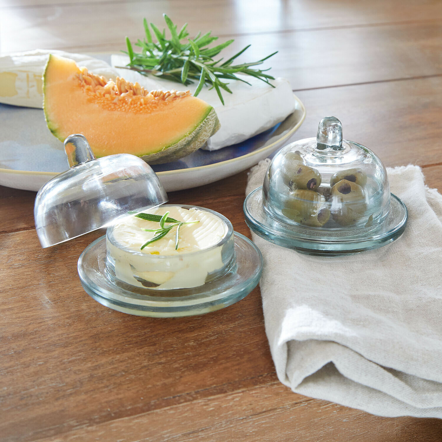 Skleněné nádoby na máslo a olivy na dřevěném stole s melounem a sýrem v pozadí