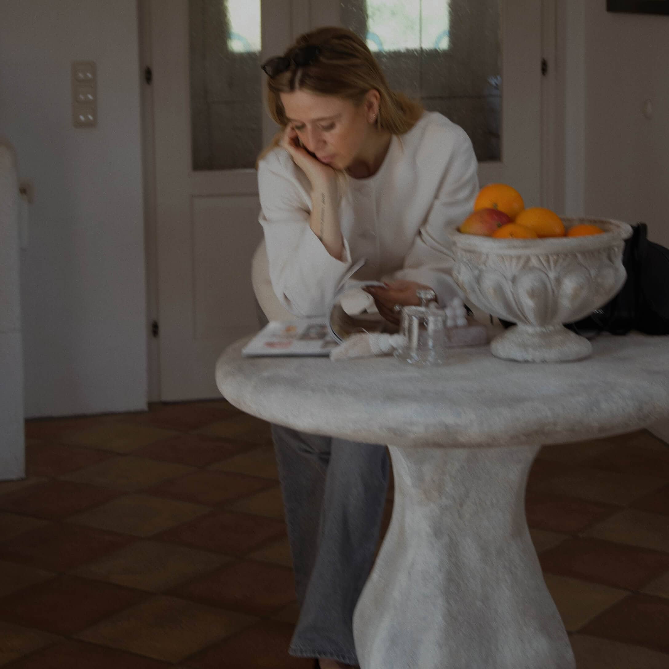 Žena v bílém svetru čte u kulatého kamenného stolu s miskou ovoce v interiéru