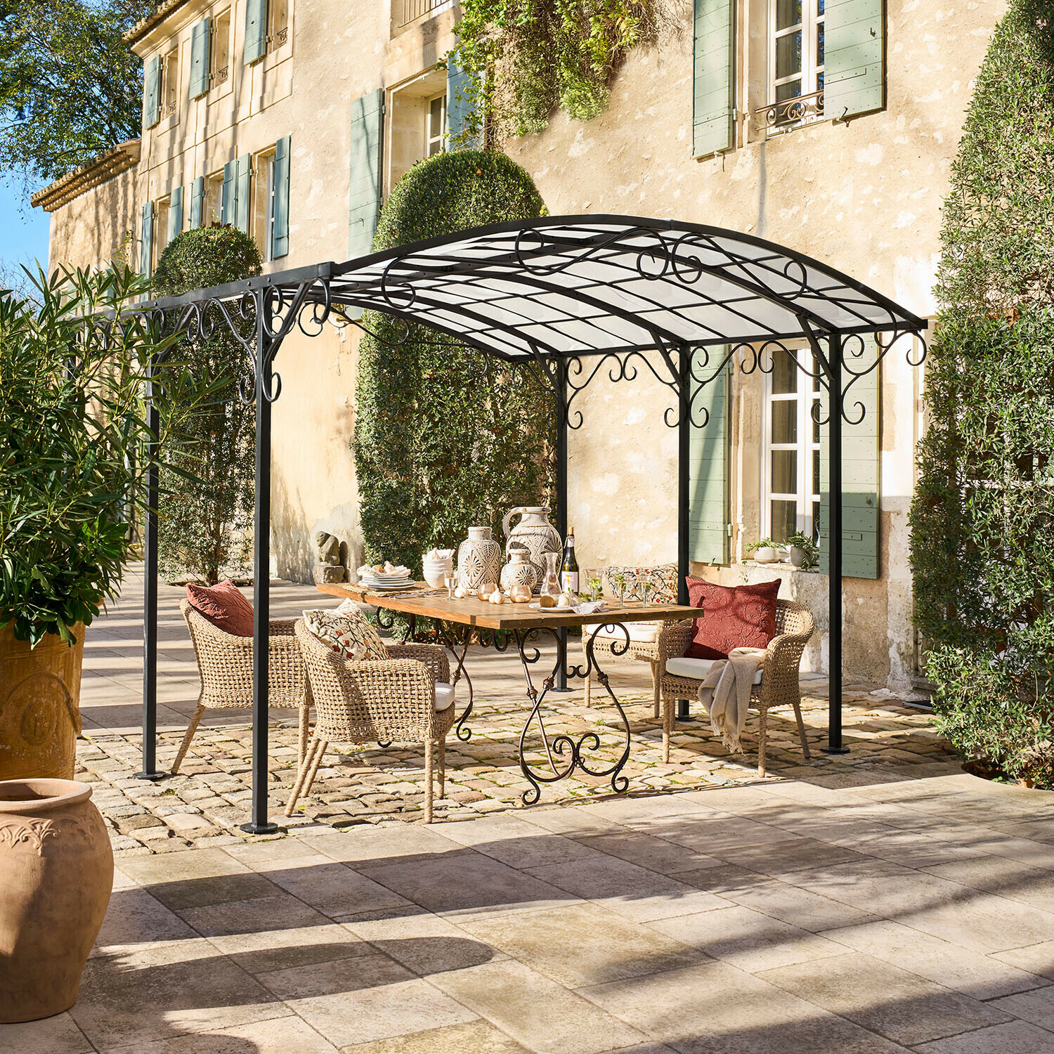 Pergola Delbridge Černá kovová zahradní pergola s ozdobnými křivkami nad dřevěným stolem a ratanovými židlemi