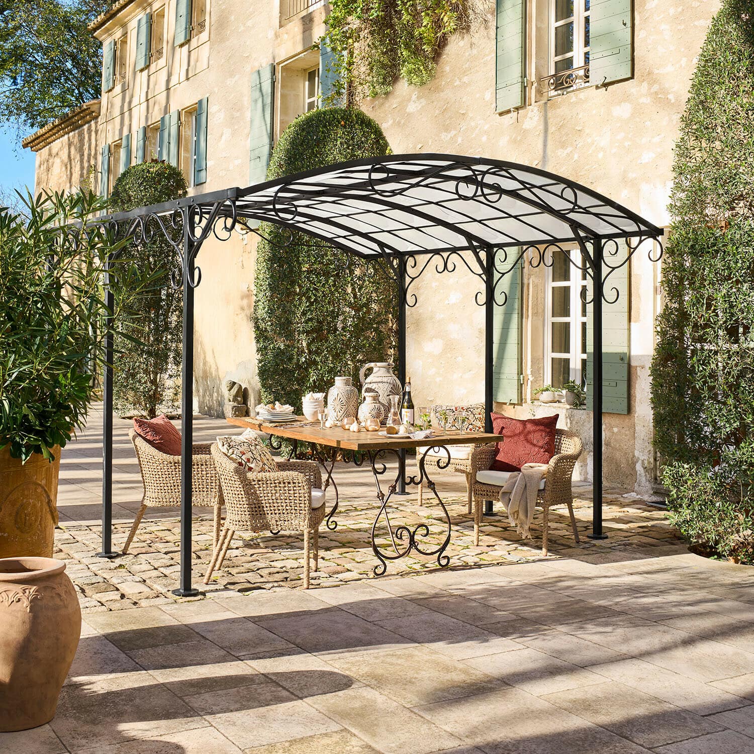 Pergola Delbridge Černá kovová zahradní pergola s ozdobnými křivkami nad dřevěným stolem a ratanovými židlemi