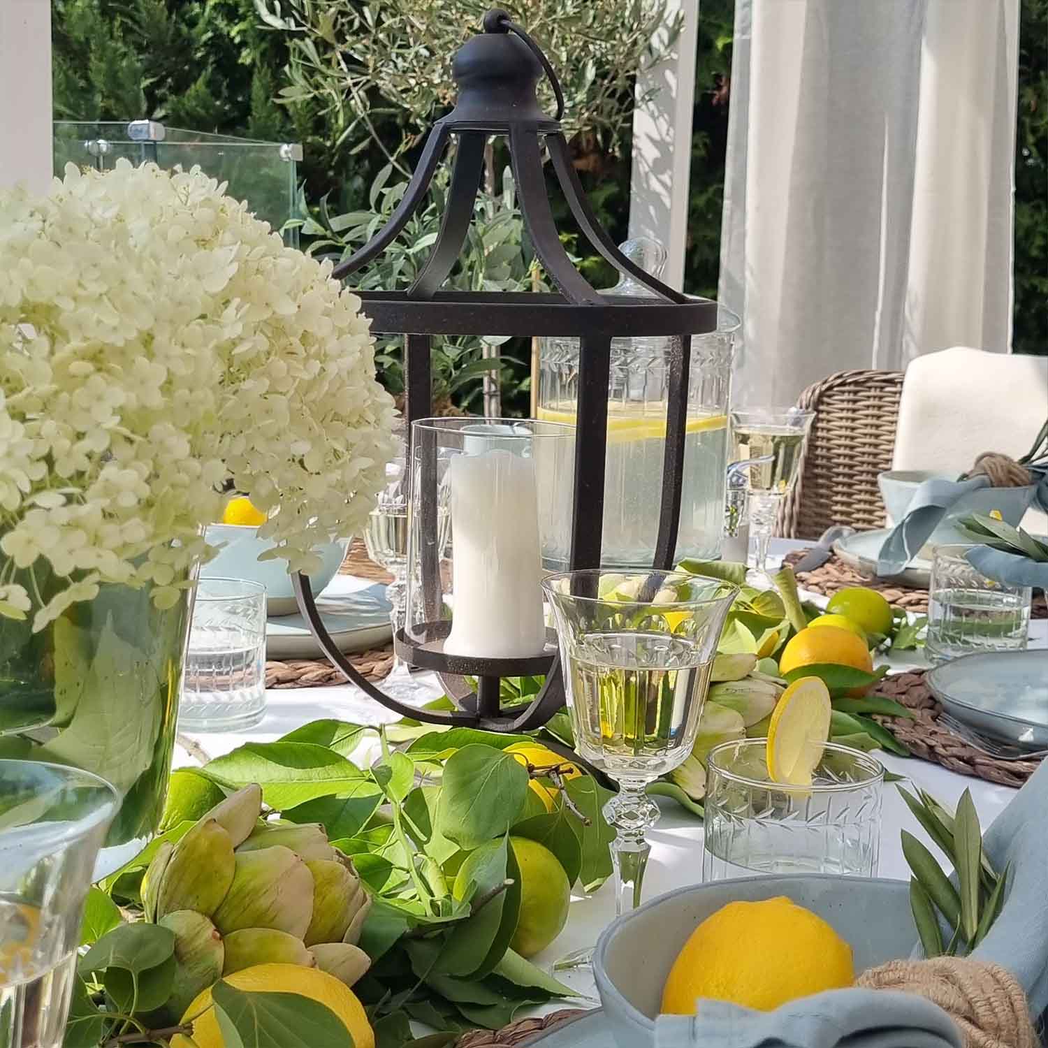 Černá kovová lucerna s bílou svíčkou na letně zdobeném stole s citrony a zelenými listy