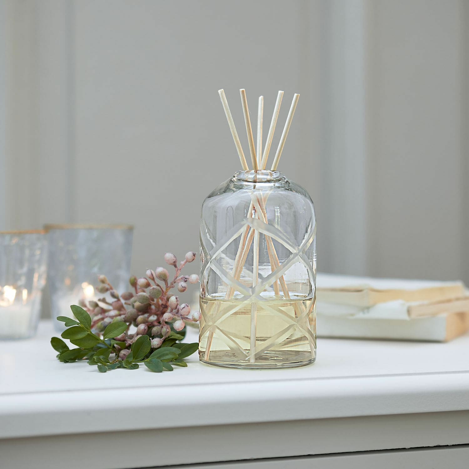 Aroma difuzér s tyčinkami Azminah Skleněný aroma difuzér s přírodními tyčinkami a žlutým olejem na bílém stole s dekorací