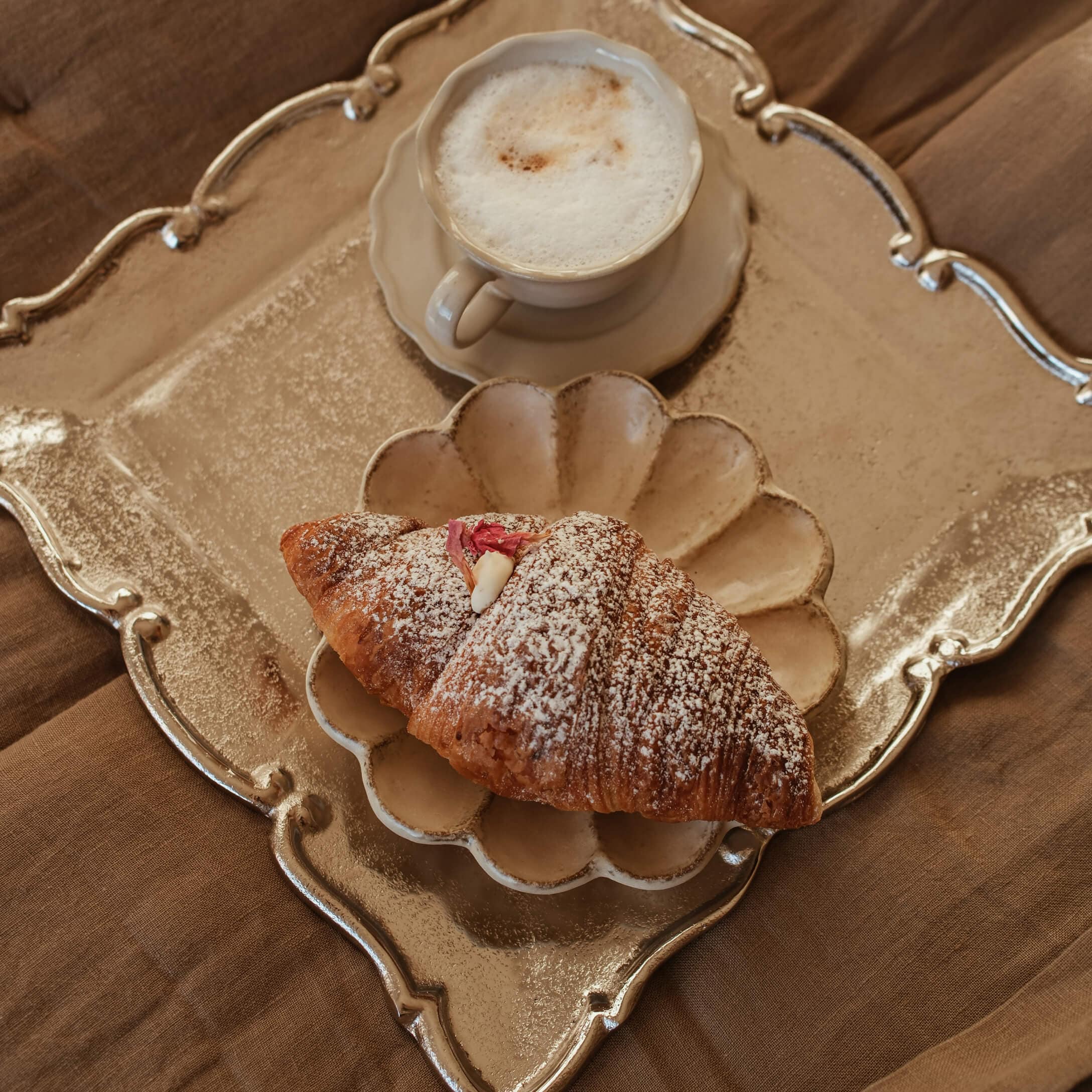 Šálek krémového cappuccina a croissant s moučkovým cukrem na béžovém talíři a zlatém podnosu