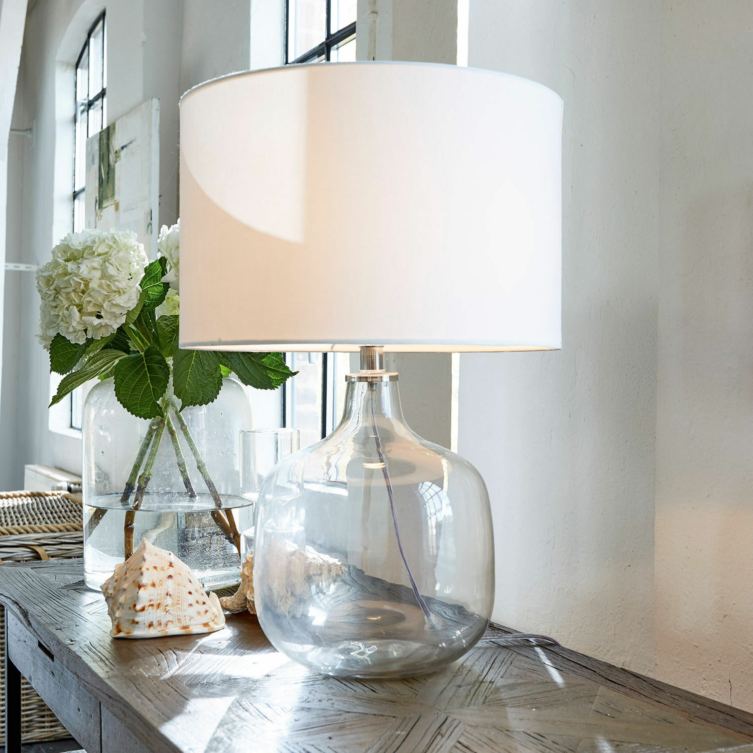 Stolní lampa Malmy Stolní lampa z průhledného skla s bílým stínidlem na dřevěném stole s dekorací mušlí