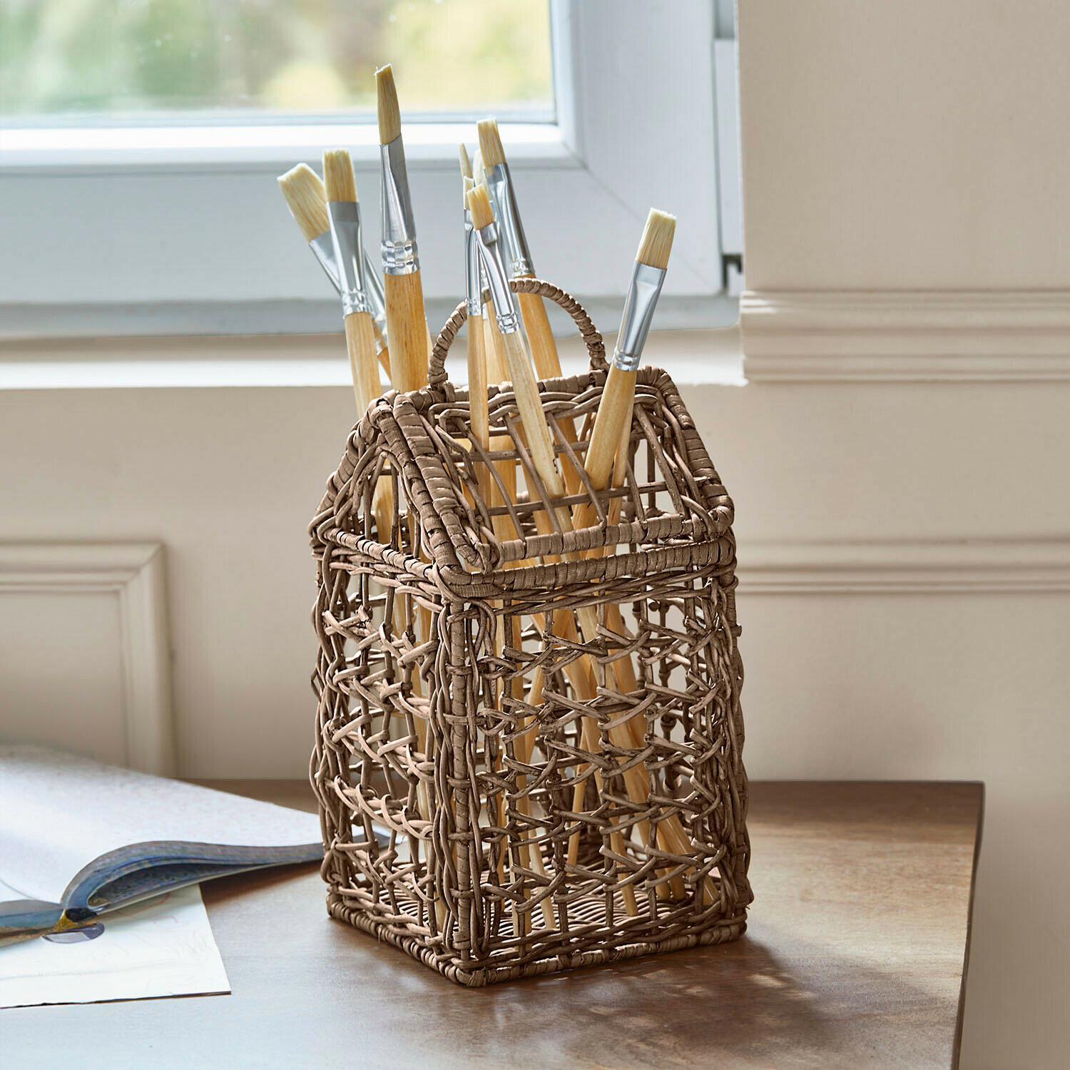 Hnědý proutěný stojan ve tvaru domu s malířskými štětci na dřevěném stole