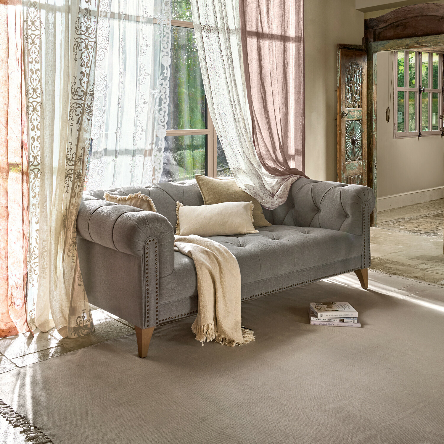 Šedá čalouněná pohovka s dřevěnými nohami a béžovými polštáři v světlém obývacím pokoji se závěsy