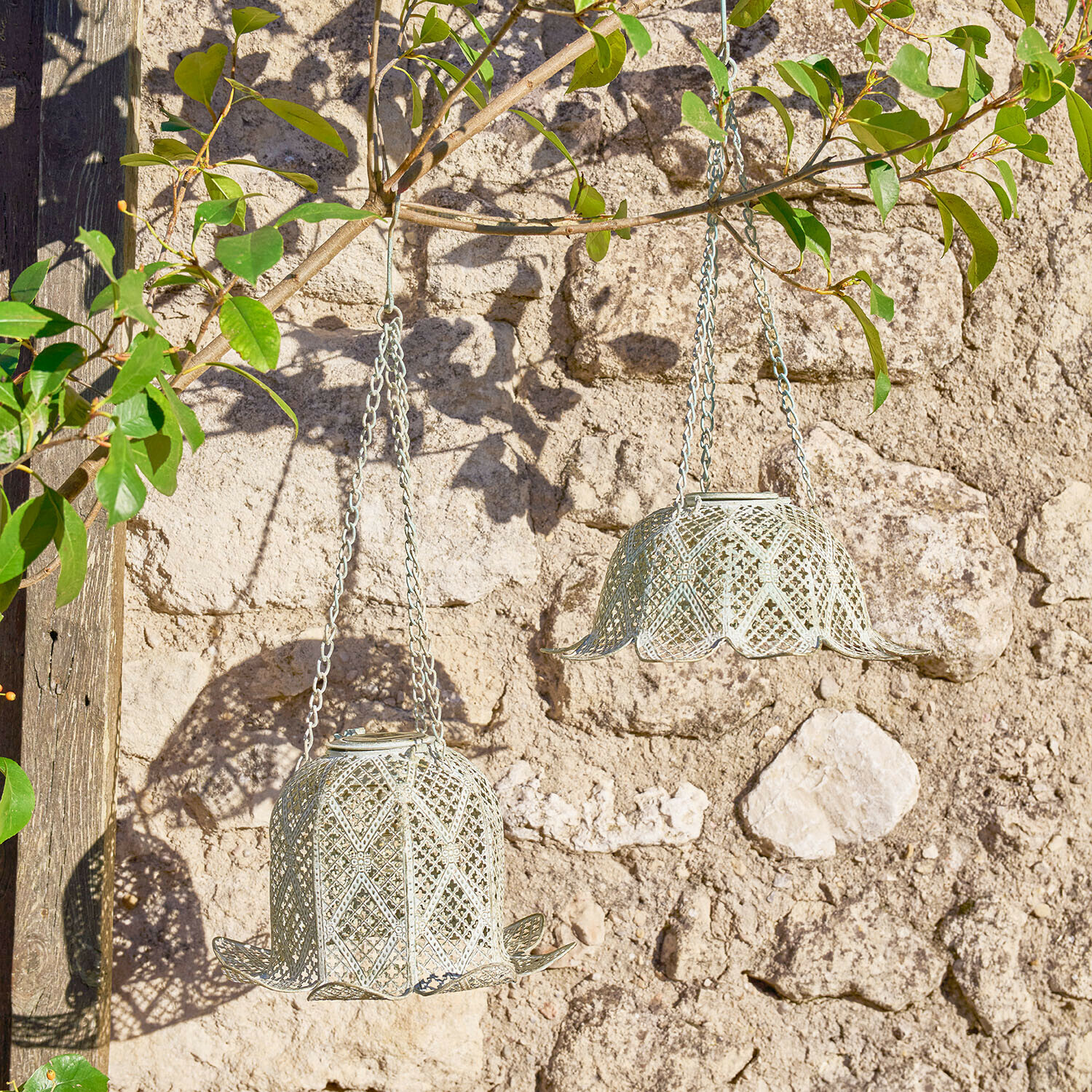 Dvě zavěšené kovové lucerny ve tvaru zvonku světle šedé barvy na kamenné zdi