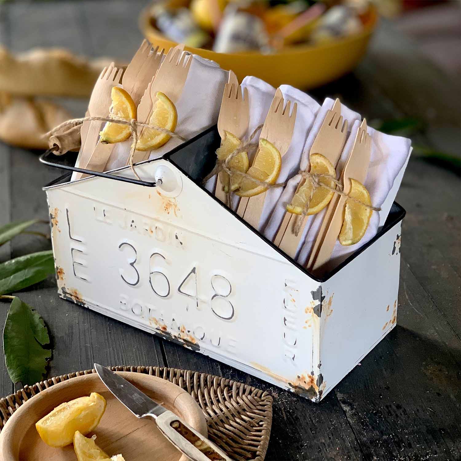 Bílý kovový box s patinou obsahující dřevěné příbory a bílé ubrousky ozdobené plátky citronu